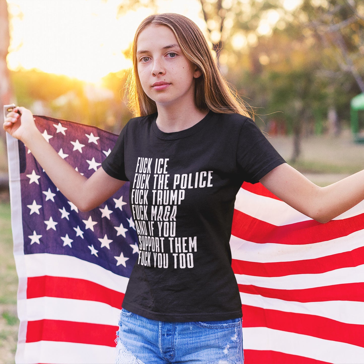 Teenage girl at sunset holding an American flag, wearing the bold Fuck 'Em All T-shirt with anti-authoritarian statements, reflecting a strong stance against oppression and supporting equality.