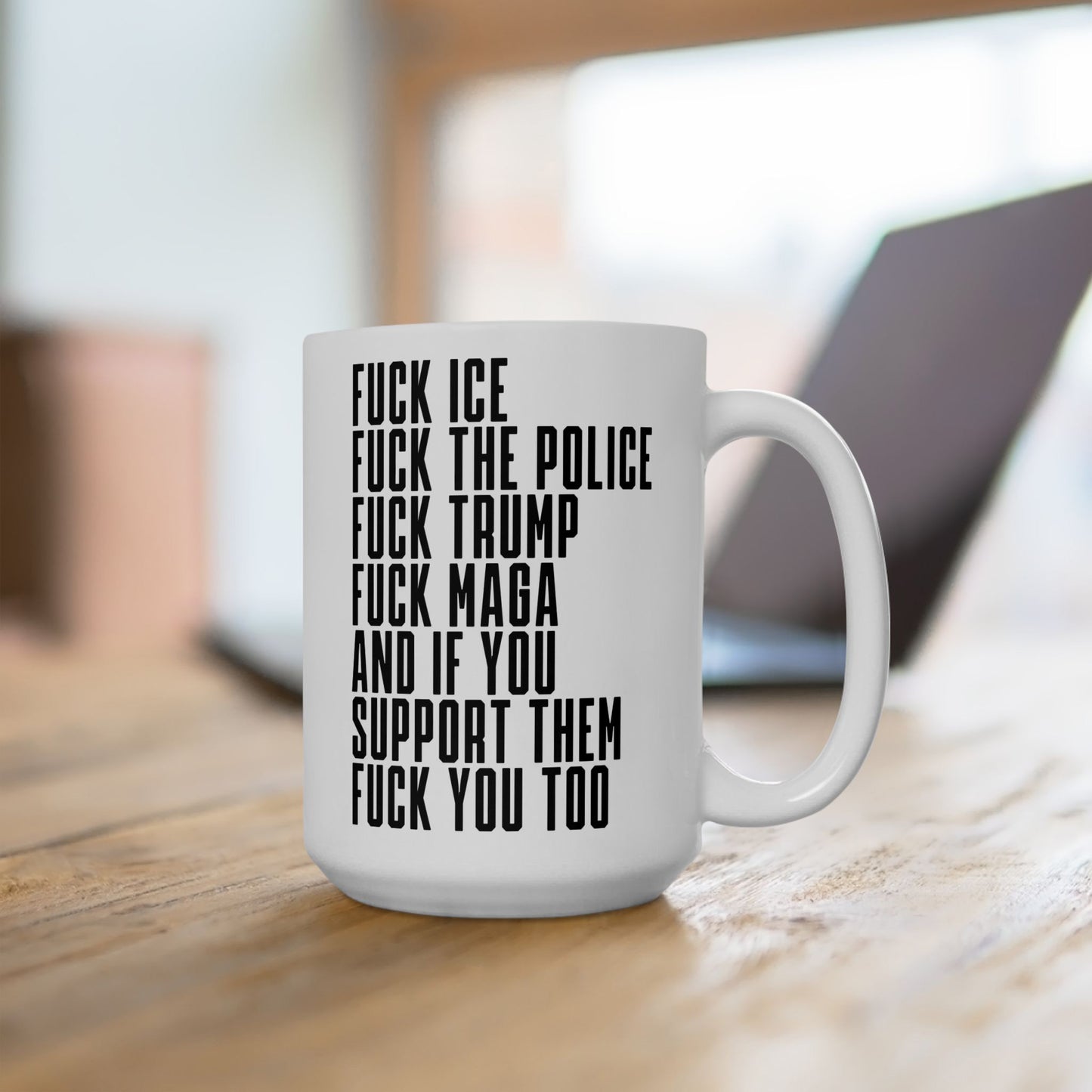 White ceramic Fuck 'Em All Mug with bold, black text listing anti-fascist and anti-establishment sentiments, sitting on a wooden surface with a blurred laptop in the background.