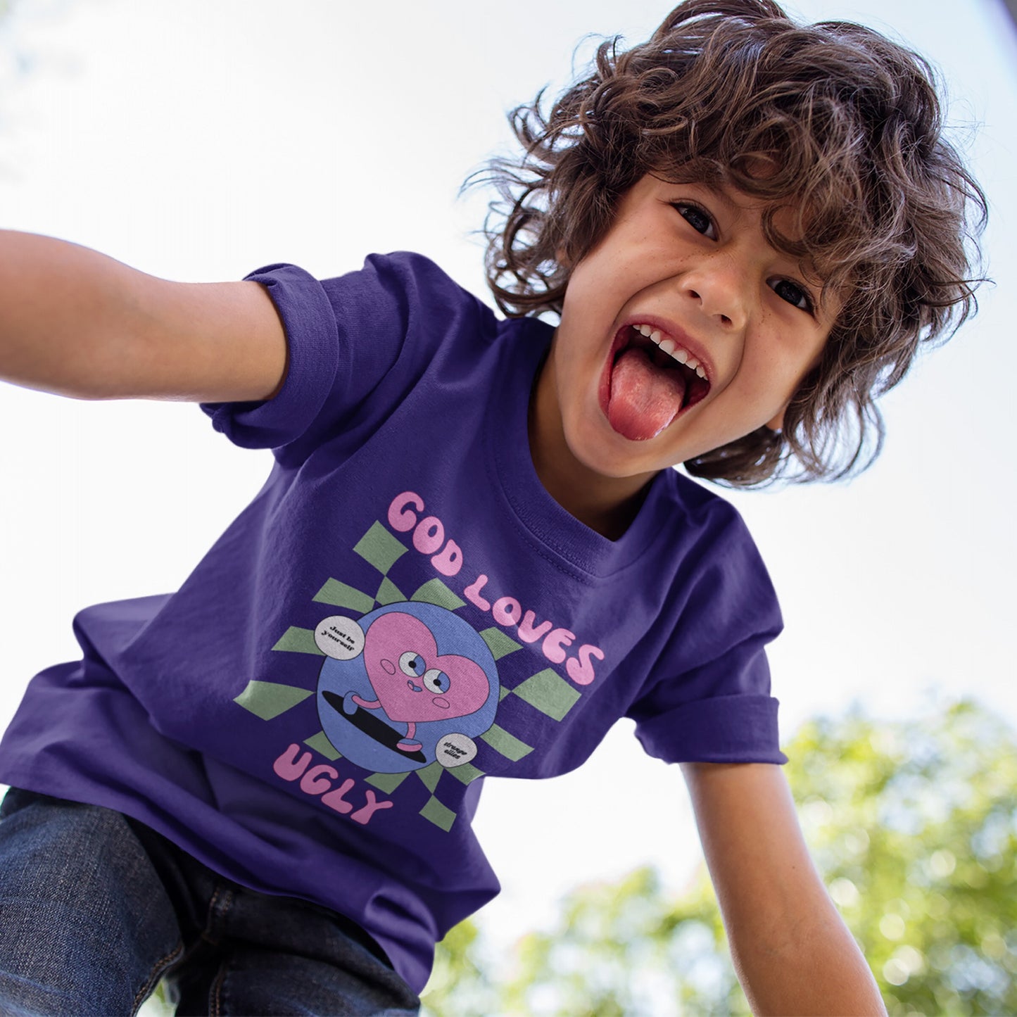 Child wearing the God Loves Ugly T-shirt featuring a funky cartoon heart with affirmations, playfully leaning toward the camera outdoors. The tee celebrates self-worth with its bold, anti-perfection message.