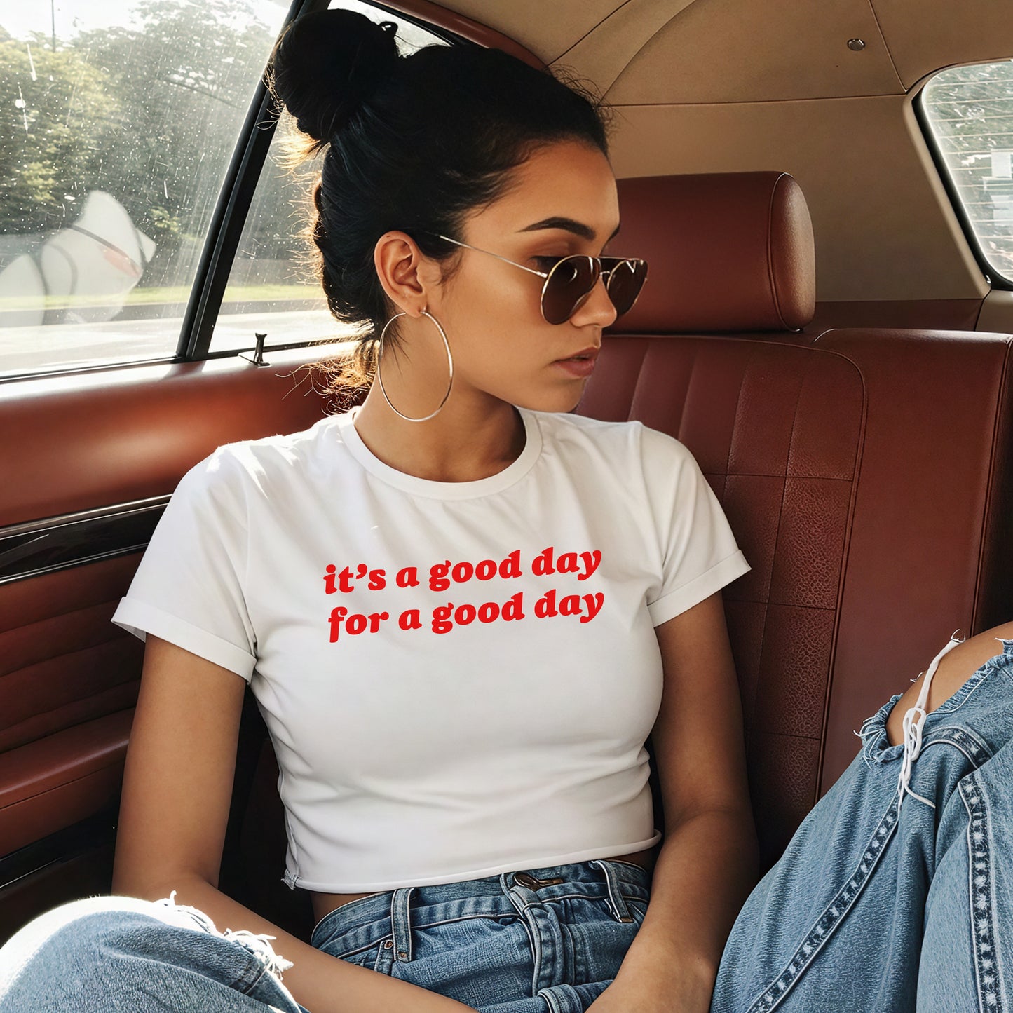 Young person in a vintage car wears the Good Day Women's Baby Tee, featuring bold red lettering it’s a good day for a good day, with hoop earrings and sunglasses, embodying retro optimism.