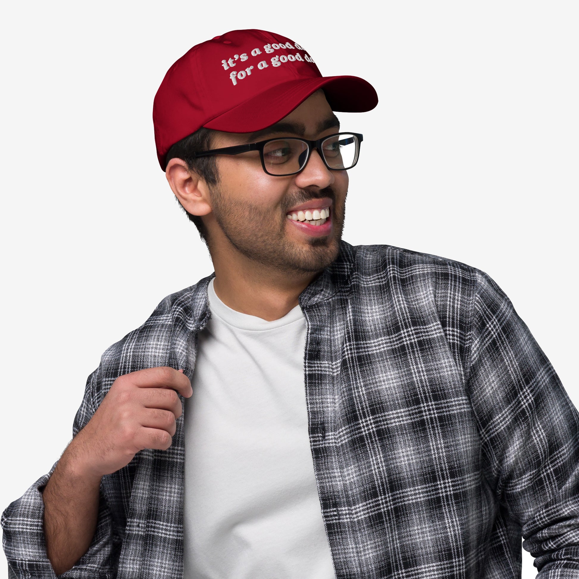 Young man wearing the Good Day Hat, a red cap with “it’s a good day for a good day” embroidered on the front, paired with glasses and a plaid shirt, smiling confidently.