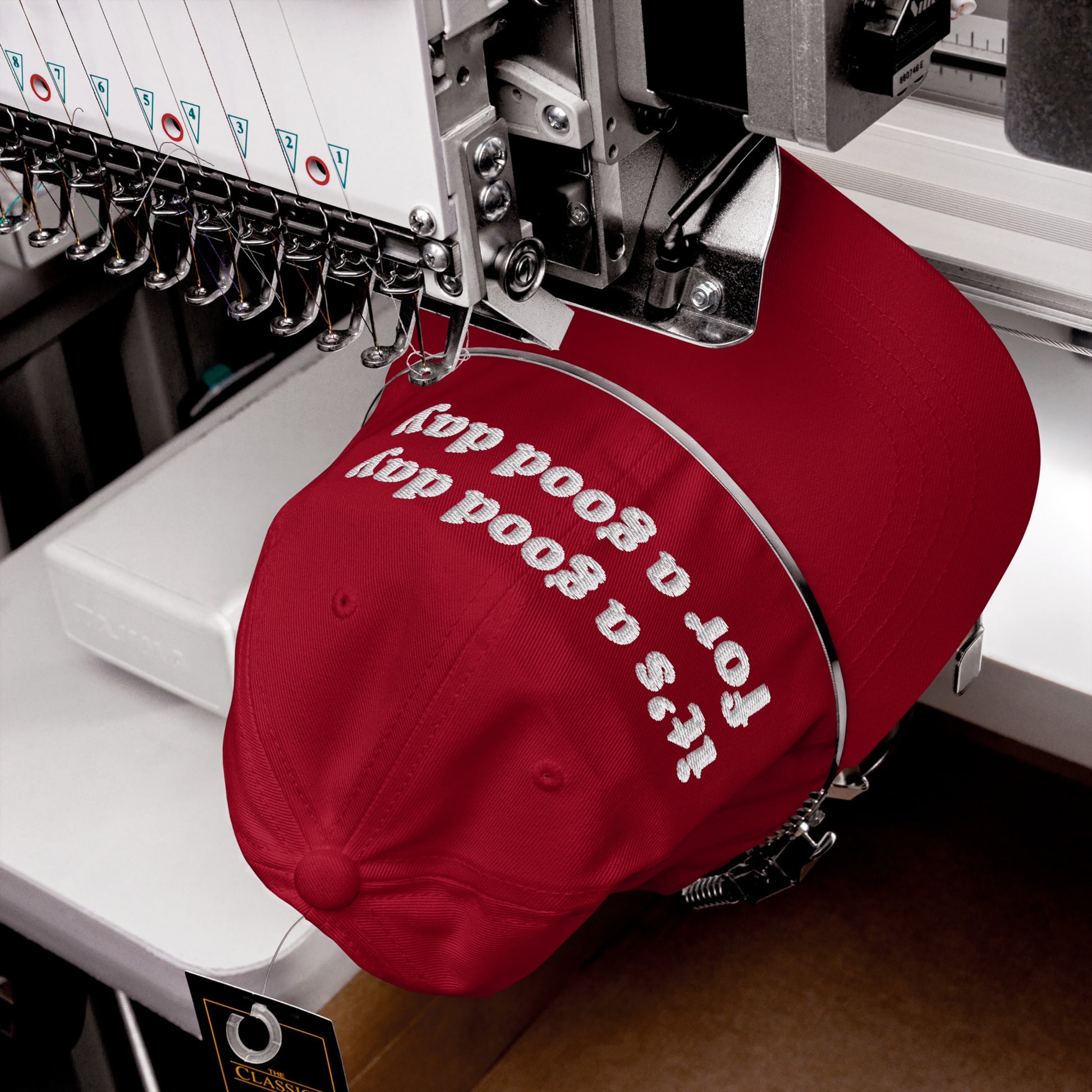 Industrial embroidery machine stitching It's a good day for a good day in white on a red baseball cap, emphasizing positivity and hopefulness with the Good Day Hat.