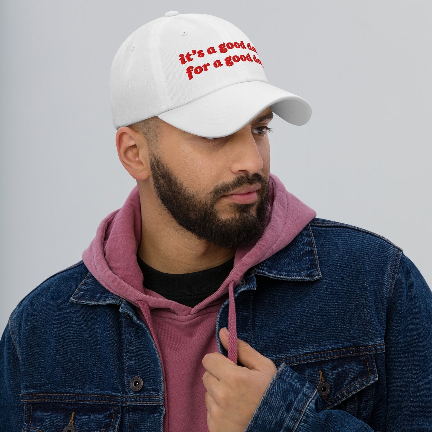Young man wearing the Good Day Hat in white with red embroidery, paired with a dusty-rose hoodie and denim jacket. The hat promotes optimism and supports Amnesty International through purchases.