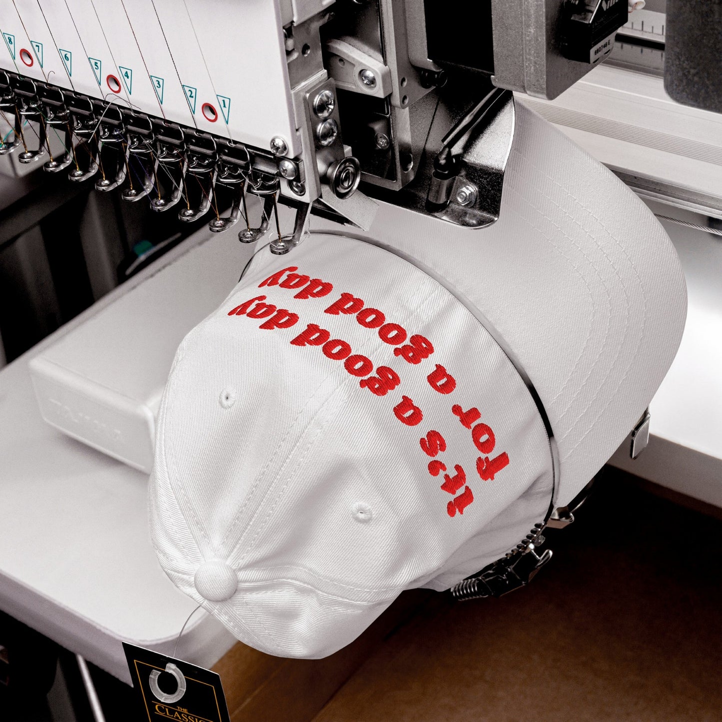 The Good Day Hat, a white cotton cap, is being embroidered with it's for a good cause on an industrial machine, reflecting a blend of retro style and optimistic advocacy.