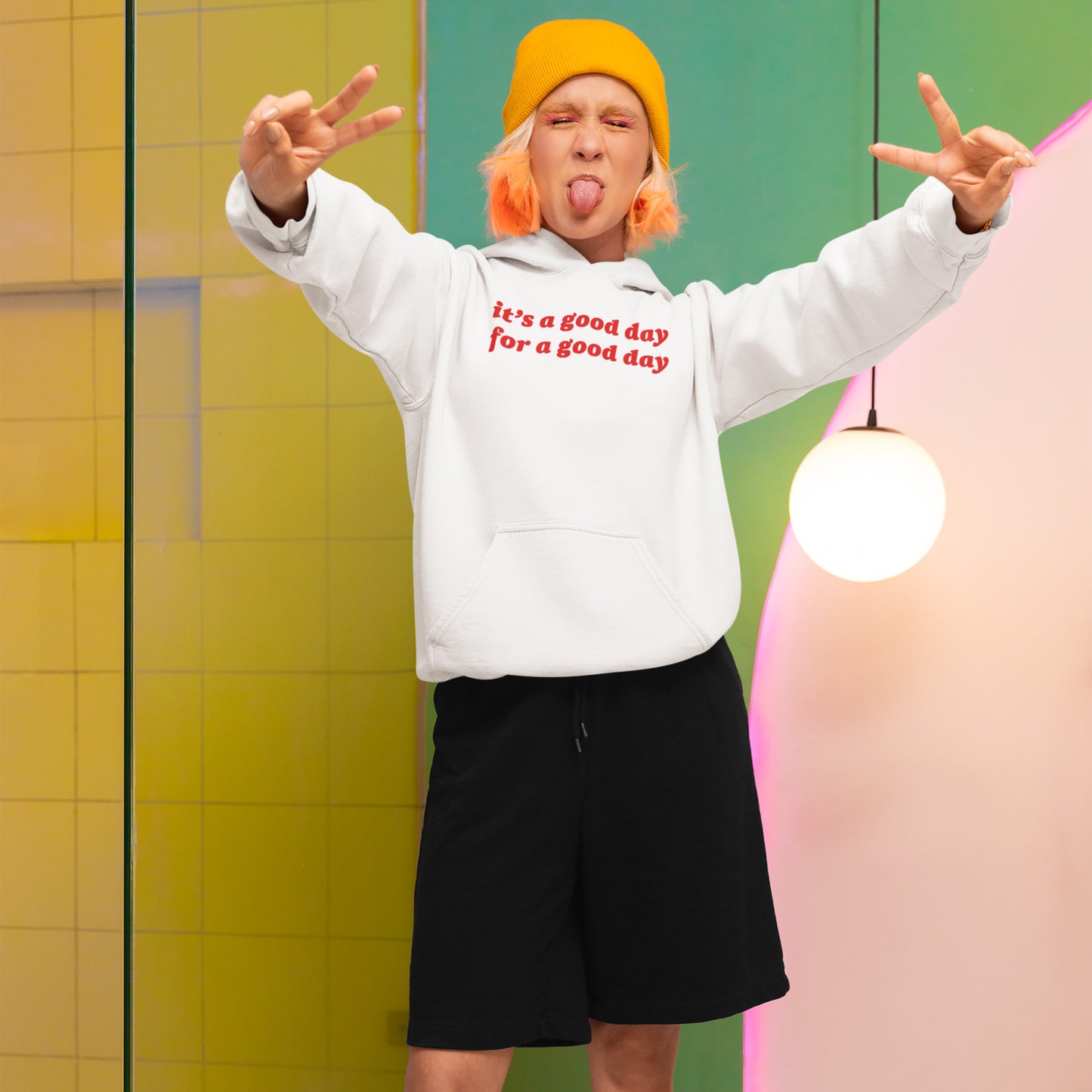 Person playfully poses wearing the Good Day Shirt hoodie, featuring red text for positivity. The vibrant setting enhances its nostalgic charm, perfect for spreading joy and supporting NAMI.