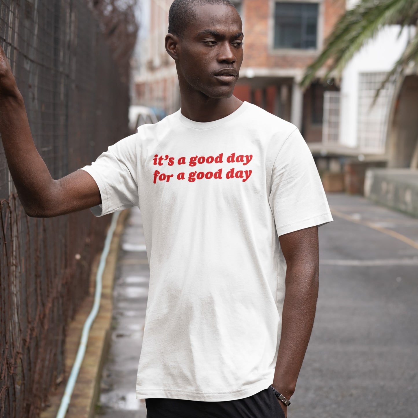 Young man leans on a fence wearing the Good Day T-shirt, featuring bold red script, “it’s a good day for a good day,” embodying optimistic vibes in a candid, urban setting.