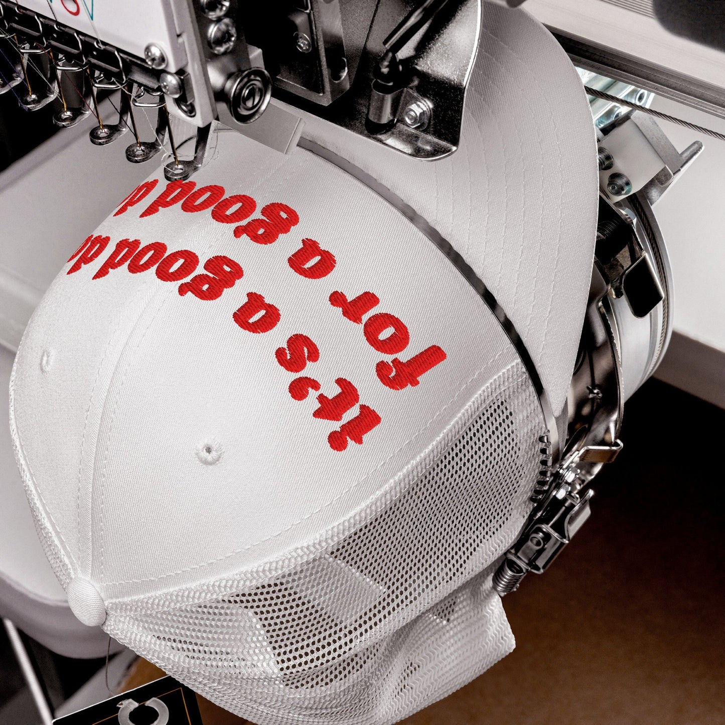 White mesh-back trucker cap titled Good Day Hat being embroidered with bold red letters on an industrial machine, embodying retro optimism and mental health advocacy, aligning with equality and support values.