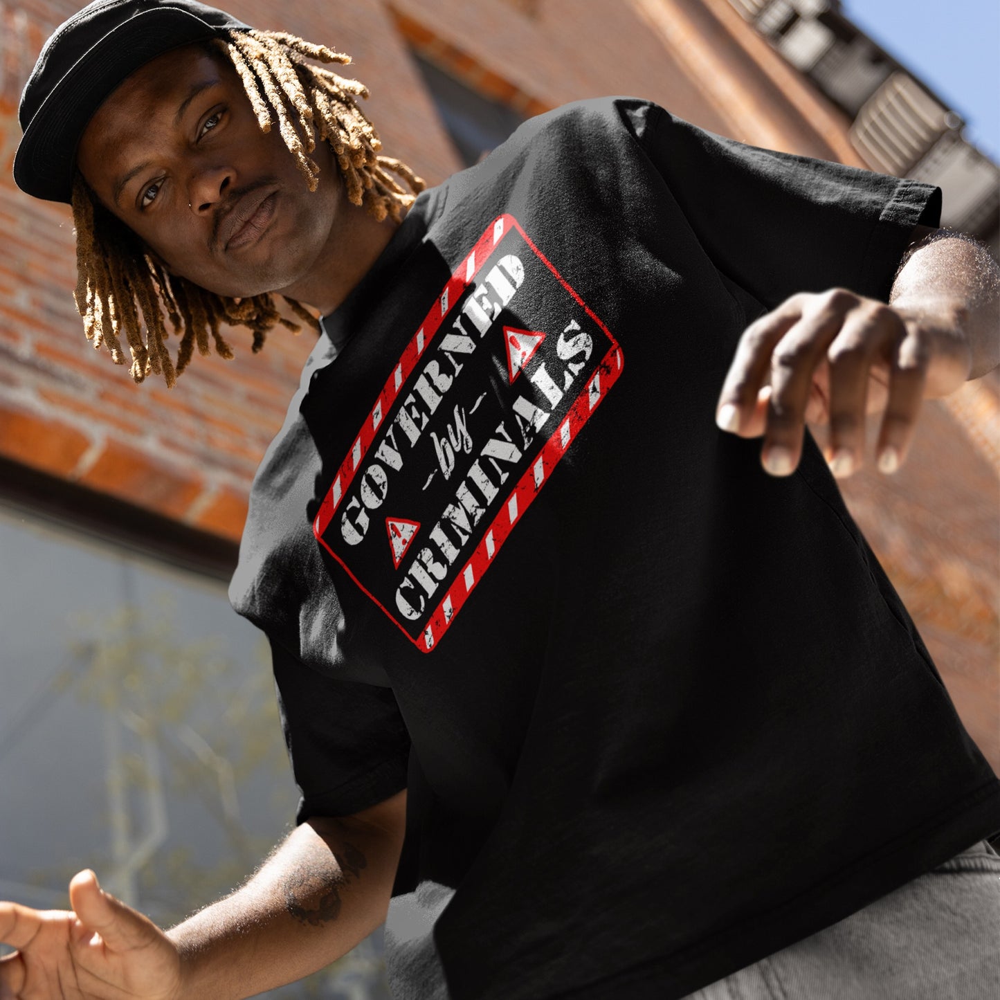 Young man in black Governed By Criminals T-shirt with red-and-white stencil design, embodying a bold statement against corruption and elite power, standing outdoors with blond dreadlocks under a cap.
