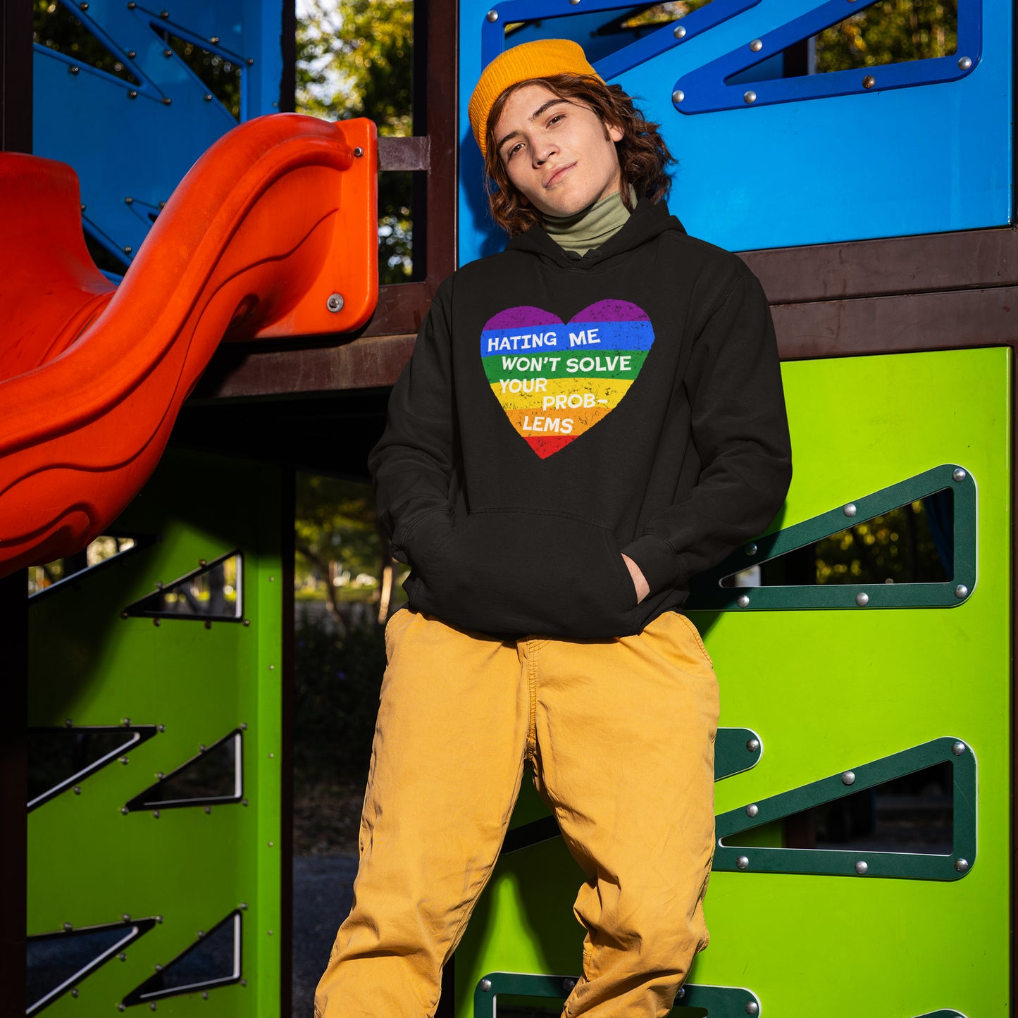 Young person leans against a playground structure wearing a No LGBTQ Hate hoodie featuring a rainbow-striped heart with the message, “HATING ME WON’T SOLVE YOUR PROBLEMS,” supporting LGBTQ pride and resistance.