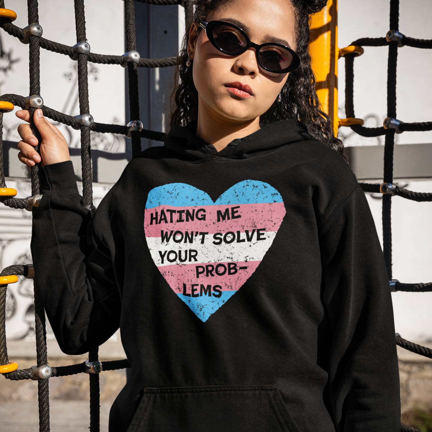 Person in black hoodie with trans flag heart and slogan HATING ME WON’T SOLVE YOUR PROB-LEMS. The No Trans Hate Shirt promotes visibility and pride, supporting The Trevor Project.