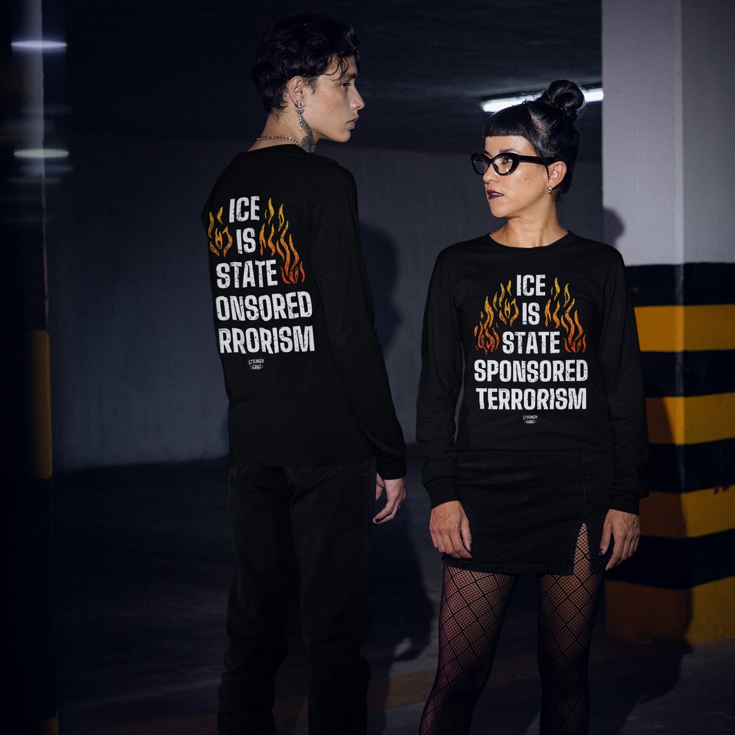 Two people wear the ICE Is State Sponsored Terrorism T-shirt featuring bold flaming designs and protest text, showcased front and back in a dimly lit parking garage.