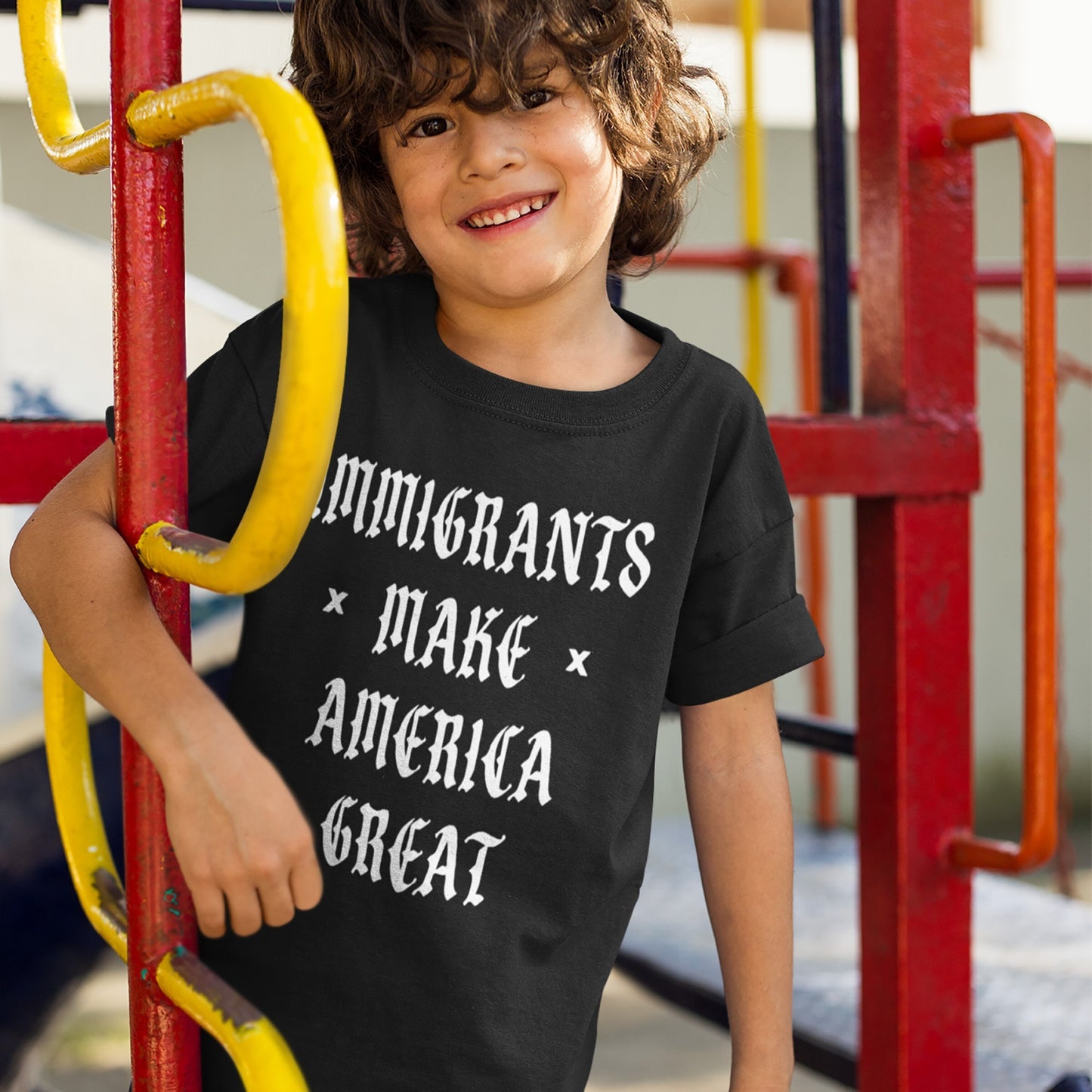 Child smiling on a jungle gym, wearing the Immigrants Make America Great T-shirt in black with bold white Gothic lettering, advocating for immigrant rights and equality.