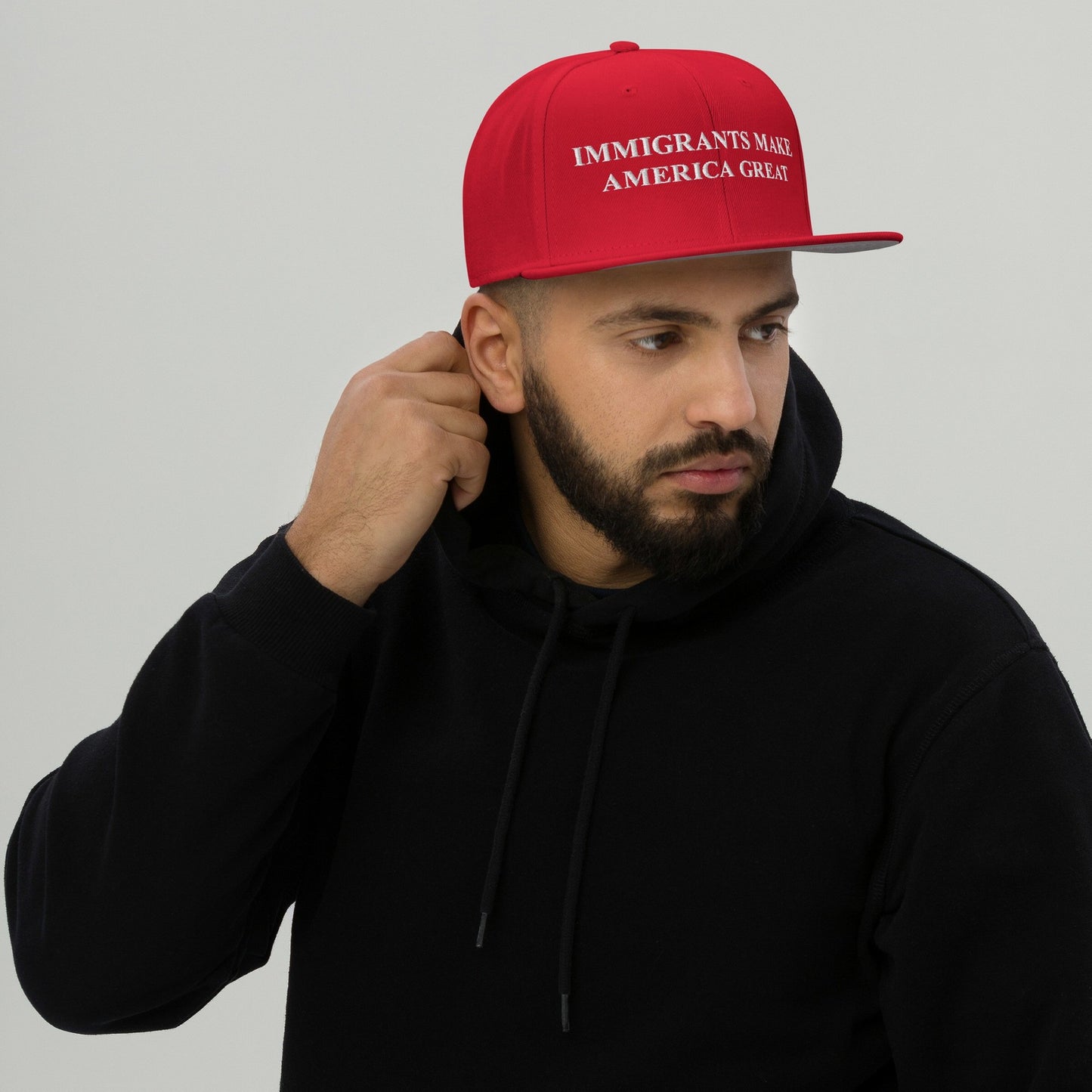 A bearded man in a black hoodie adjusts the Immigrants Make America Great Hat, featuring bold white embroidery on a red cap, emphasizing a strong pro-immigrant message.