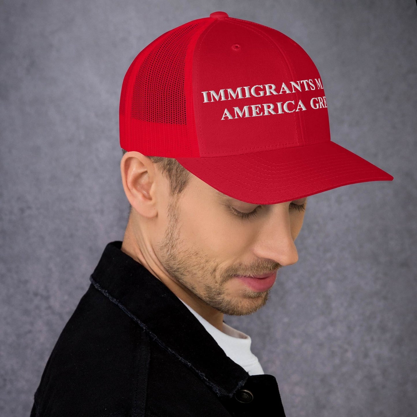 Young man in profile wears the Immigrants Make America Great Hat in red mesh-back style, featuring bold white embroidered text. His attire includes a black jacket over a white shirt against a neutral background.