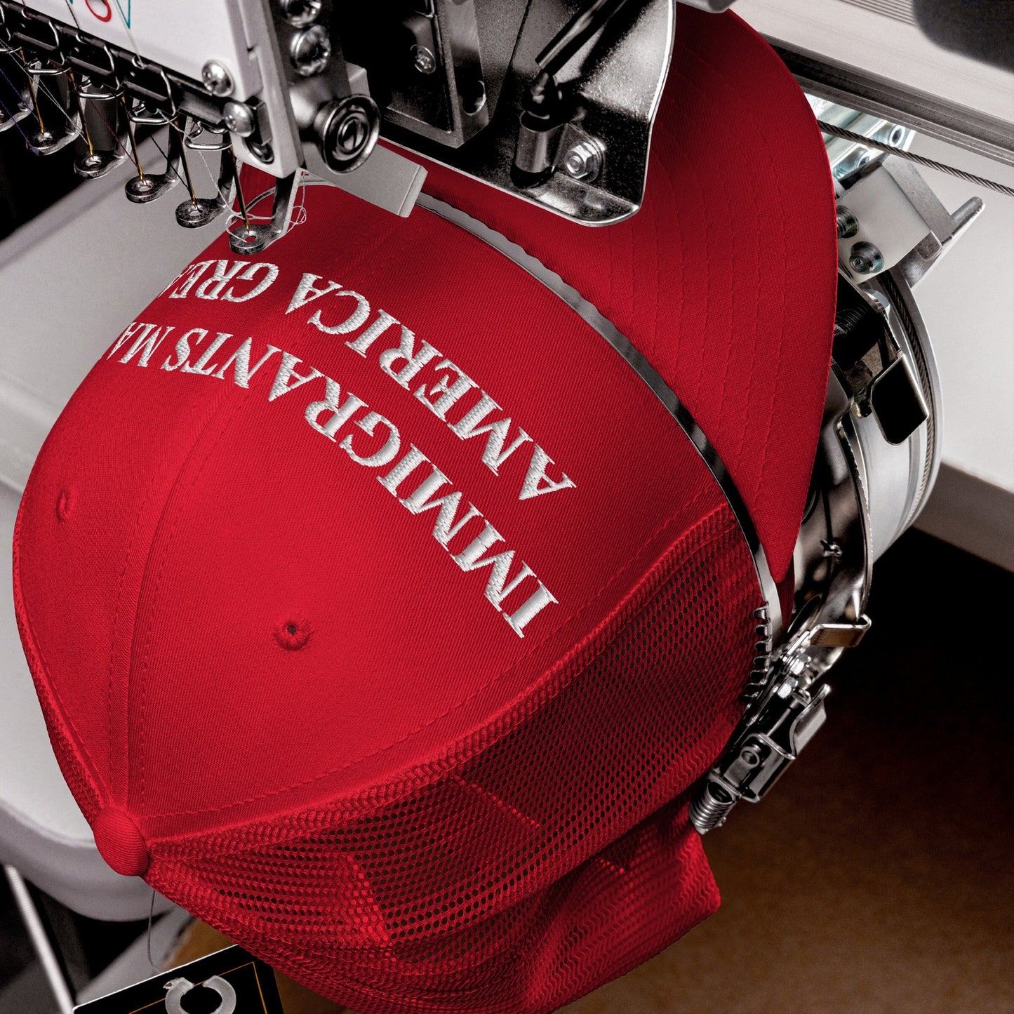 Immigrants Make America Great Hat on an embroidery machine, stitching white text onto a red mesh-back baseball cap, symbolizing solidarity and resistance against xenophobia.