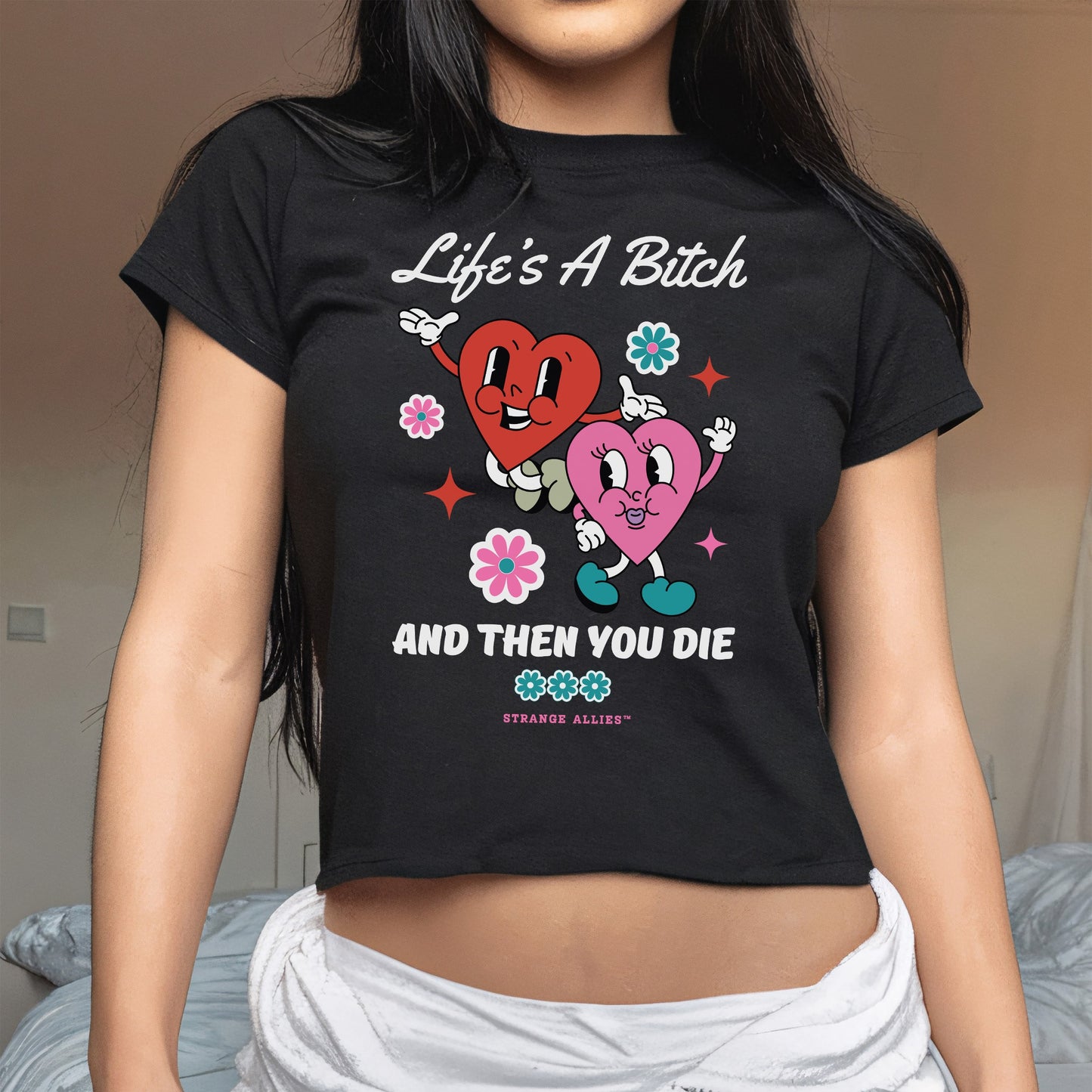 Life's A Bitch Women's Baby Tee with cartoon hearts and florals, humorously highlighting life's realities. Perfect for those embracing candid expressions and style, reflecting a blend of playful art and bold messages.