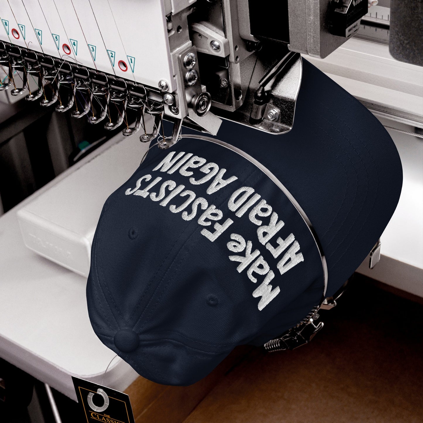 A navy-blue Make Fascists Afraid Again hat is being embroidered with white thread in bold, messy letters, showcasing the slogan on the front, held by an embroidery machine hoop.