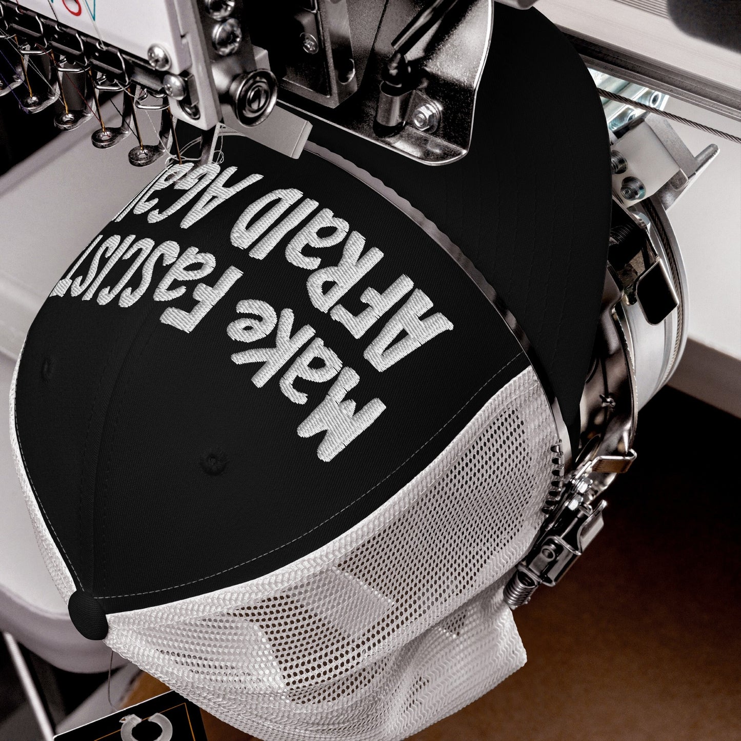 Make Fascists Afraid Again Hat in embroidery machine, white thread stitching bold text on black‐and‐white mesh trucker cap, embodying unapologetic resistance with a hand-drawn feel, ideal for active protest or daily defiance.