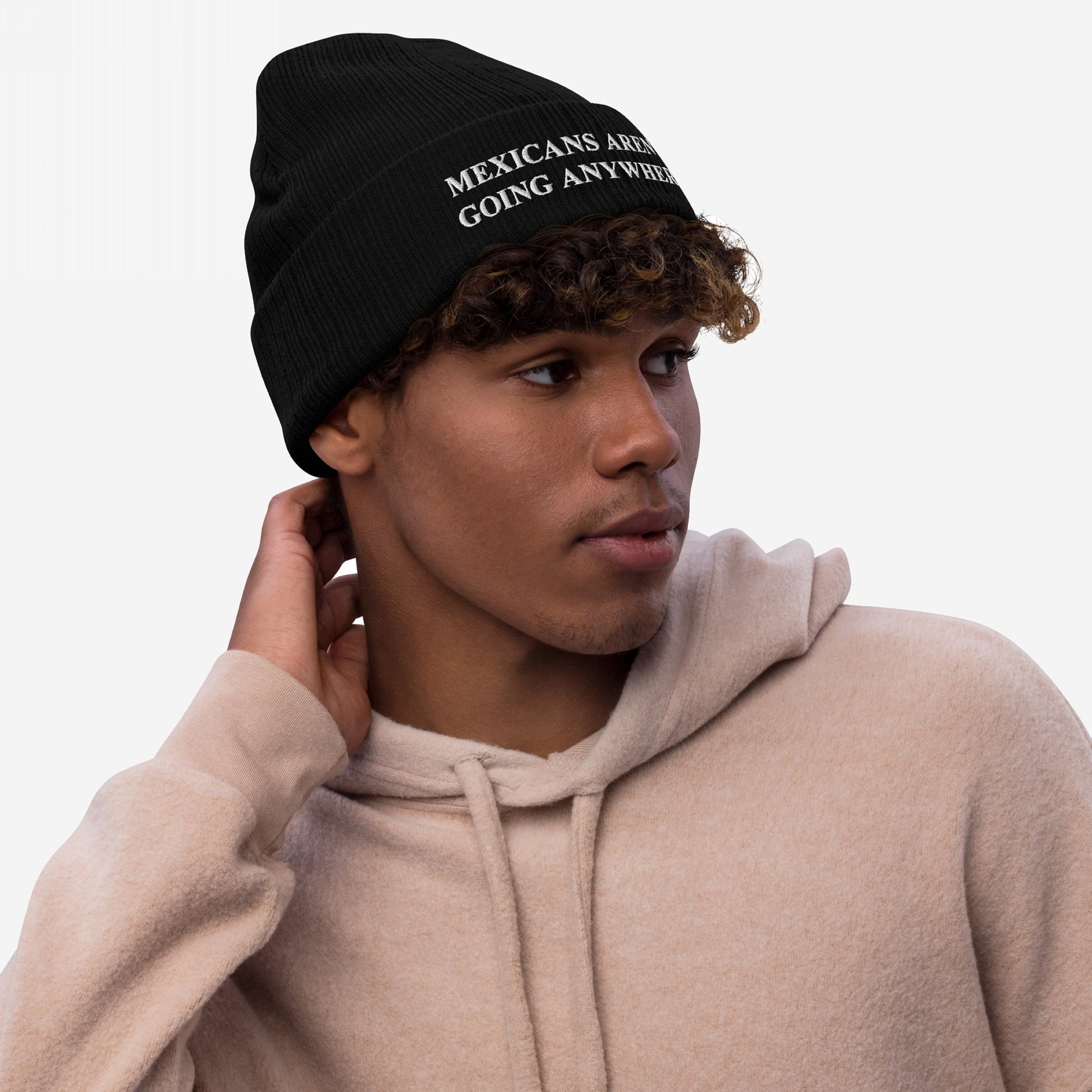 Young man wearing the Mexicans Aren’t Going Anywhere Hat, a black knit beanie with an embroidered message promoting anti-racism and immigrant pride, paired with a light-beige hooded sweatshirt.