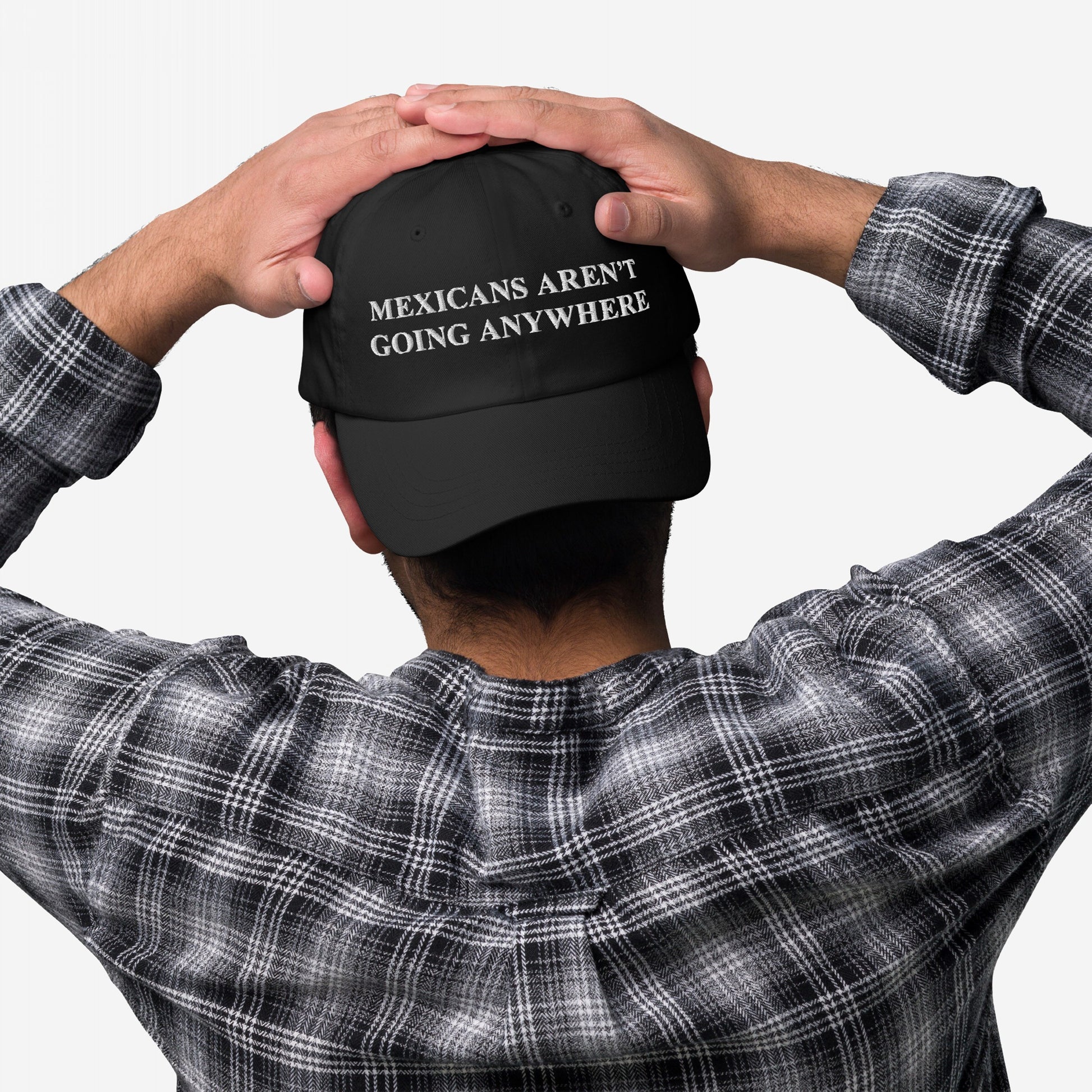 Person wearing a black Mexicans Aren’t Going Anywhere Hat with white embroidered text, paired with a gray-and-black plaid shirt, hands resting on the cap, symbolizing pride and resistance.