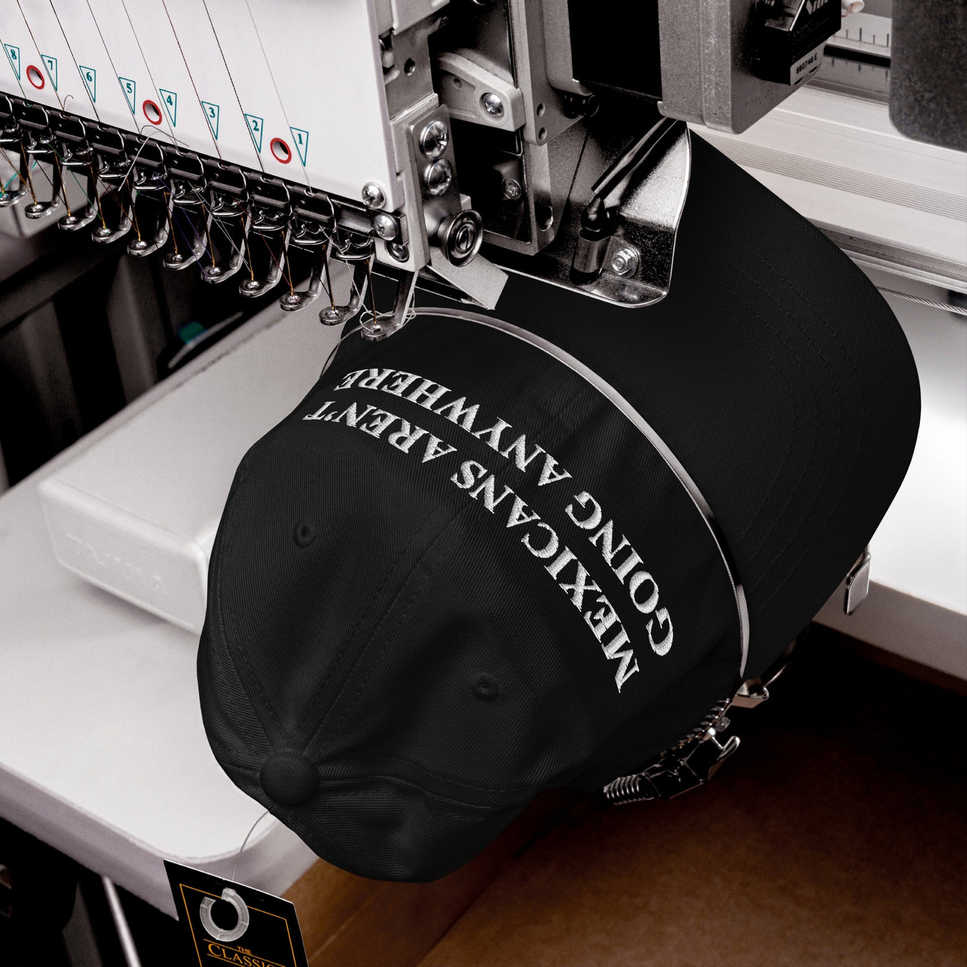 Industrial embroidery machine stitching the message “Mexicans Aren’t Going Anywhere” onto a black baseball cap, highlighting pride and resistance. Attached swing tag indicates new, classic-style headwear.
