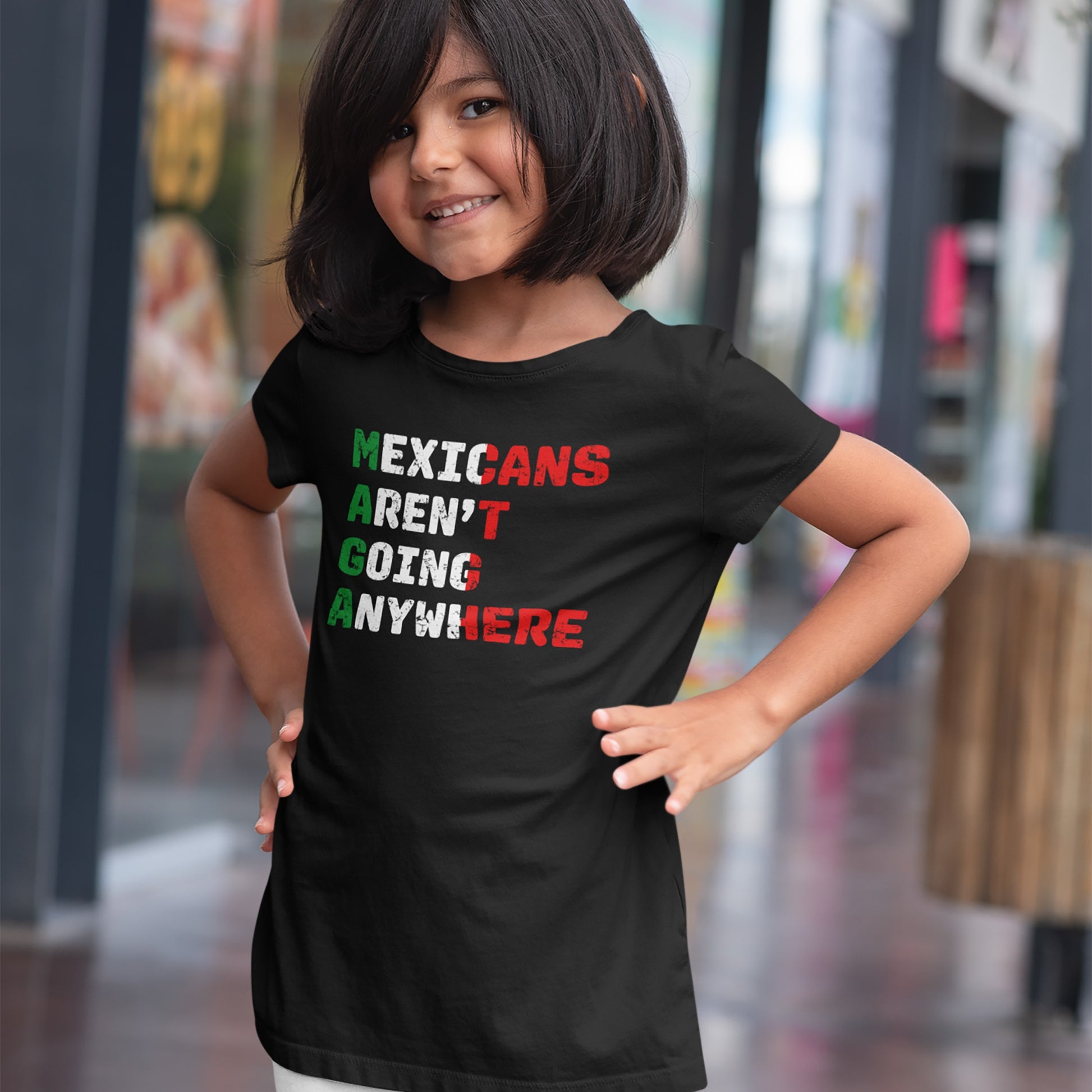 Young girl confidently poses indoors wearing the Mexicans Aren’t Going Anywhere Shirt, featuring a bold political message in red, white, and green, symbolizing resistance and pride.