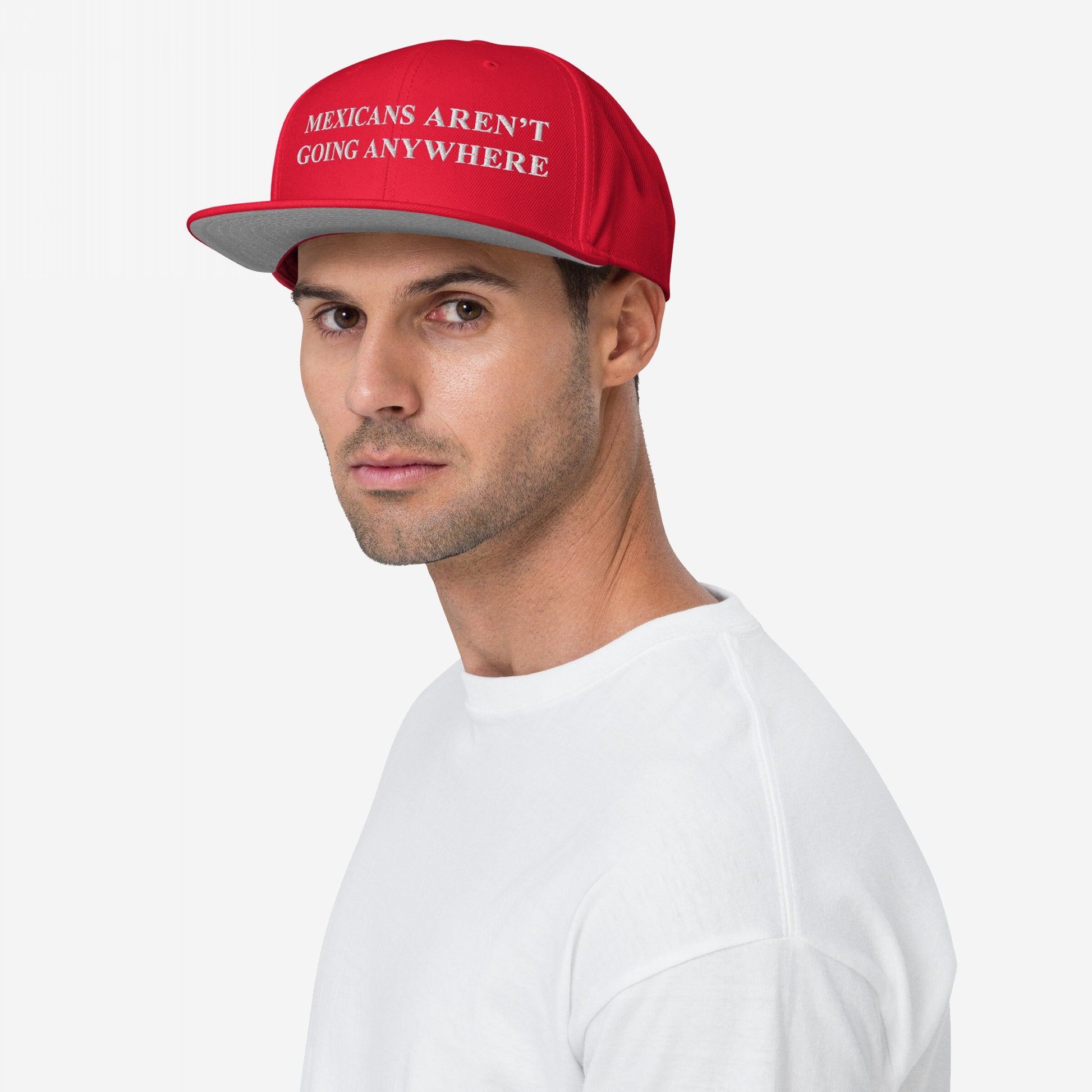Mexicans Aren’t Going Anywhere Hat: A young man in a white T-shirt wears a red cap with the slogan embroidered, symbolizing pride and resistance, set against a neutral backdrop.