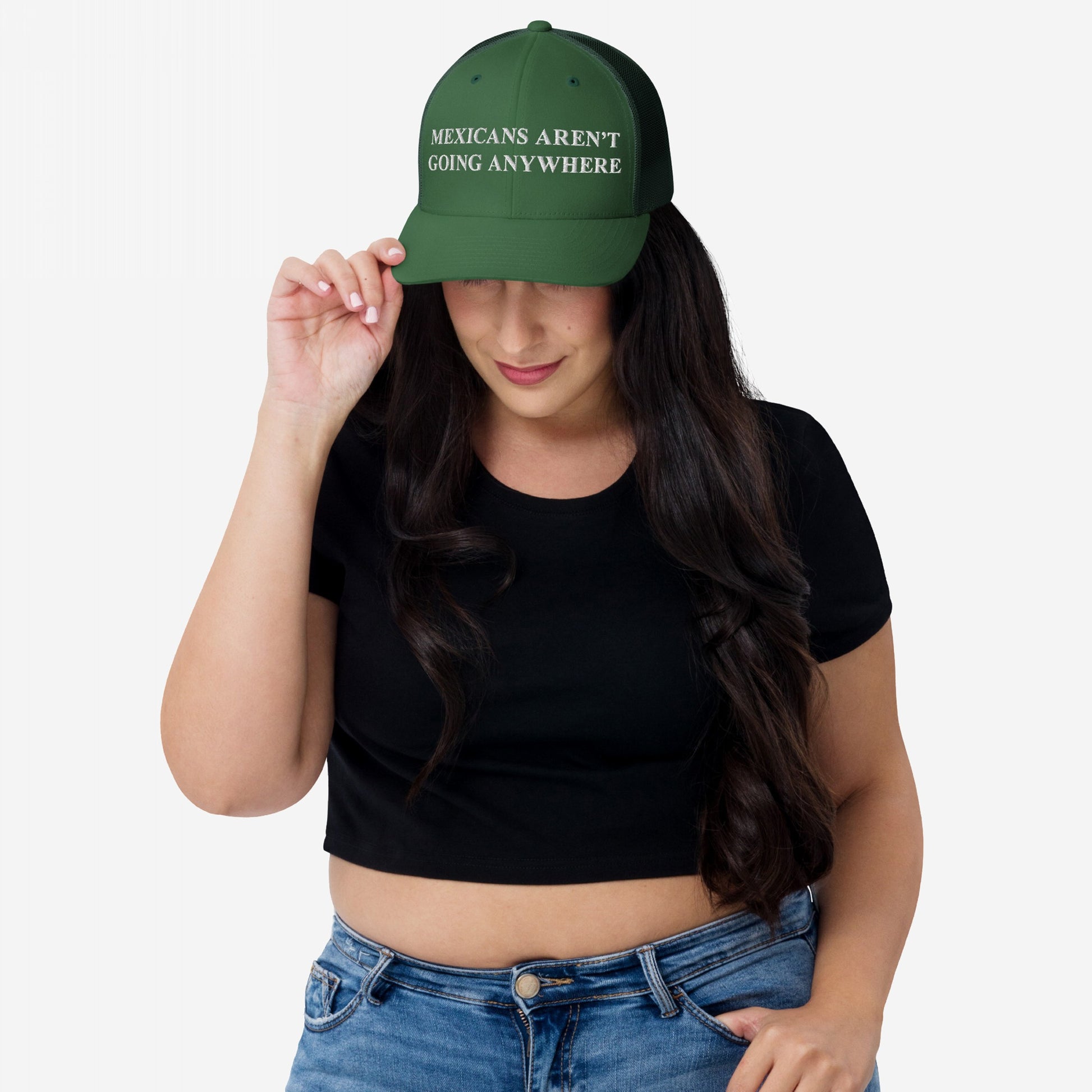 Person wearing a Mexicans Aren’t Going Anywhere dark green trucker hat, symbolizing pride and resistance. The cap features white embroidered text, with the brim slightly tipped down, complementing their black cropped T-shirt.