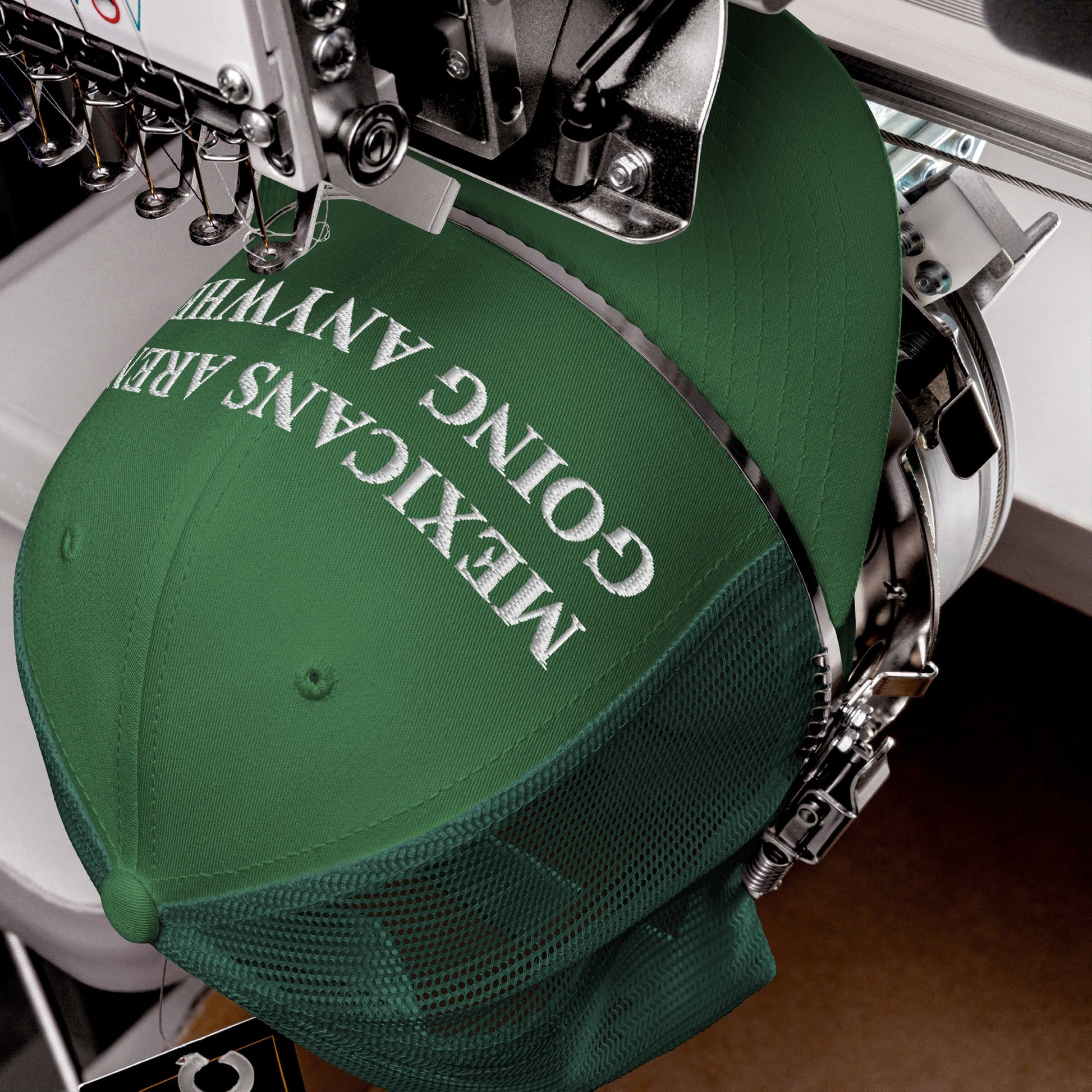 Industrial embroidery machine stitching Mexicans Aren’t Going Anywhere onto a green mesh-back baseball cap, highlighting resistance and pride. The white letters are being carefully embroidered onto the cap's front panel.