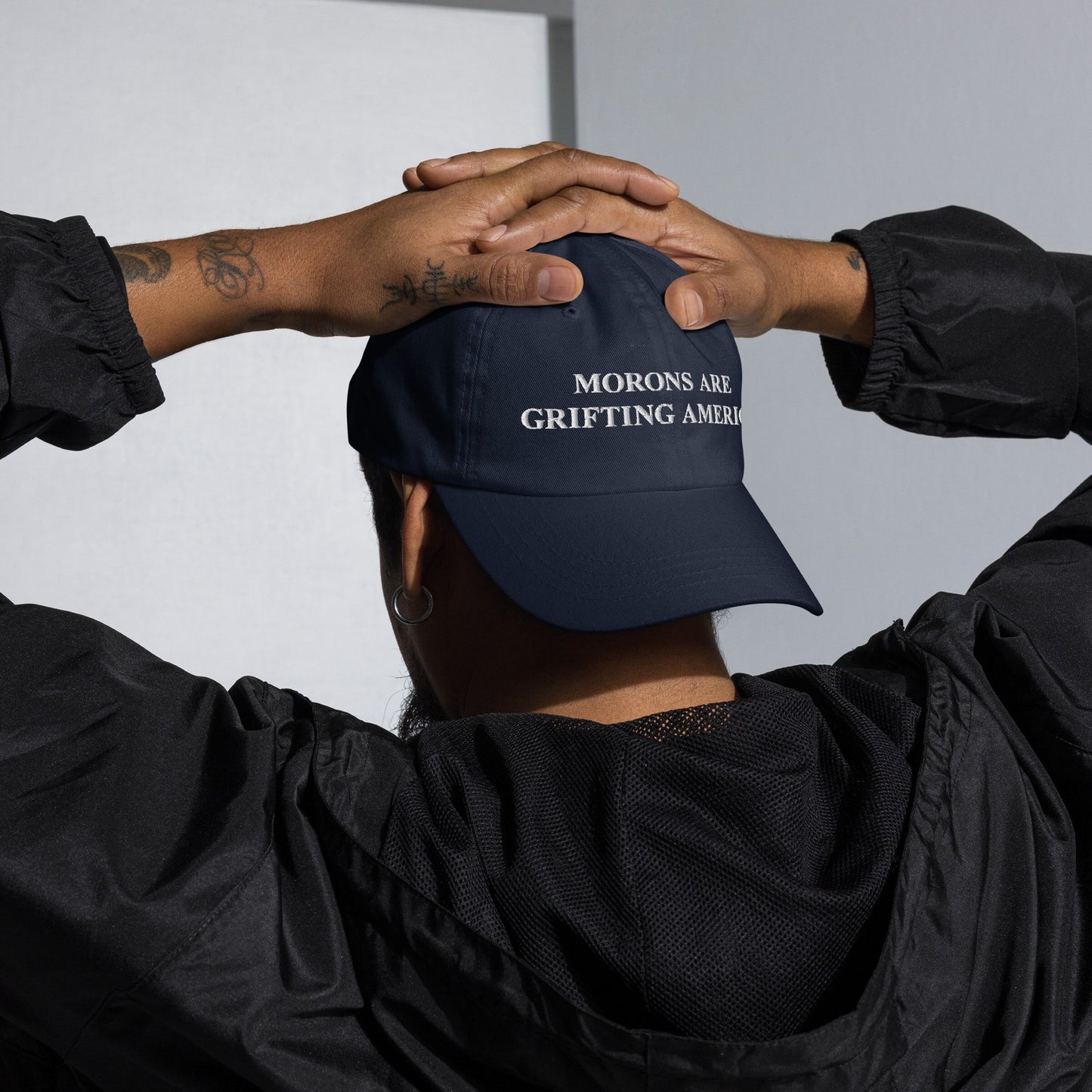 Person from behind wearing the Morons Are Grifting America hat, embroidered in white. Both hands on head, showing tattoos and earring, reflecting a bold political statement against societal norms.