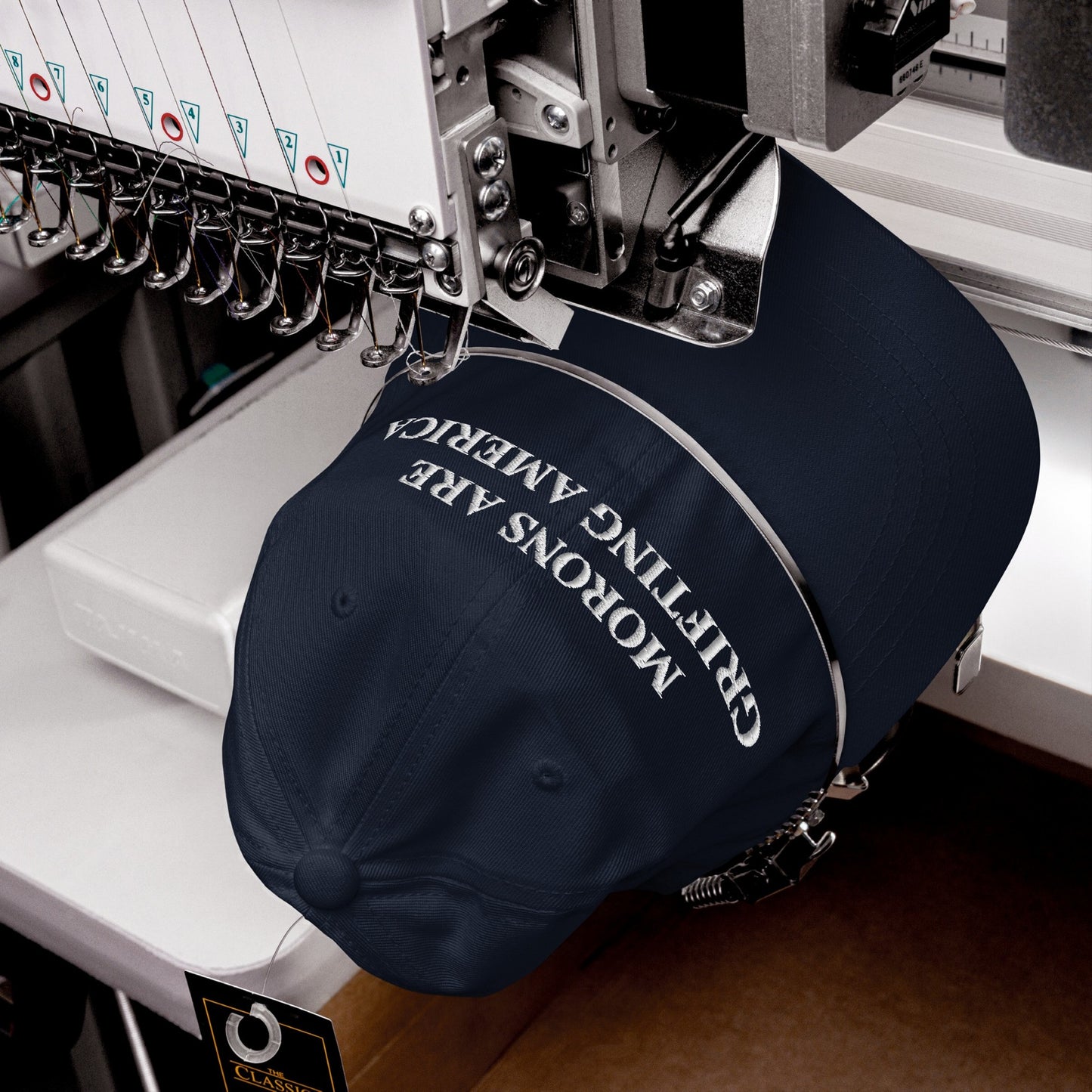 Industrial embroidery machine stitches Morons Are Grifting America in white thread on a navy baseball cap with a swing tag, reflecting a bold political statement on a snapback hat.