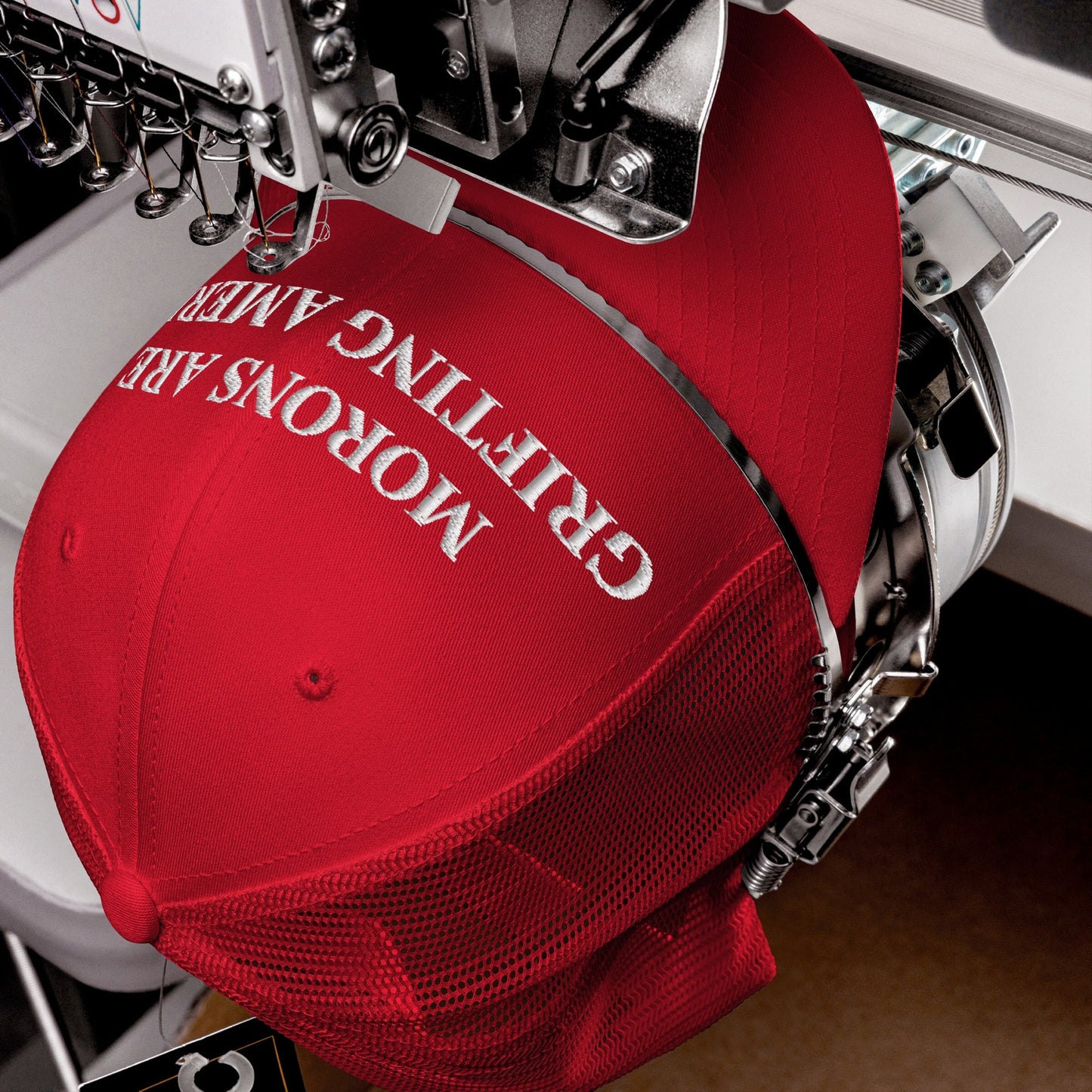 Close-up of the Morons Are Grifting America Hat on an embroidery machine, with white thread stitching bold letters on the front of a red trucker-style cap with a mesh back.