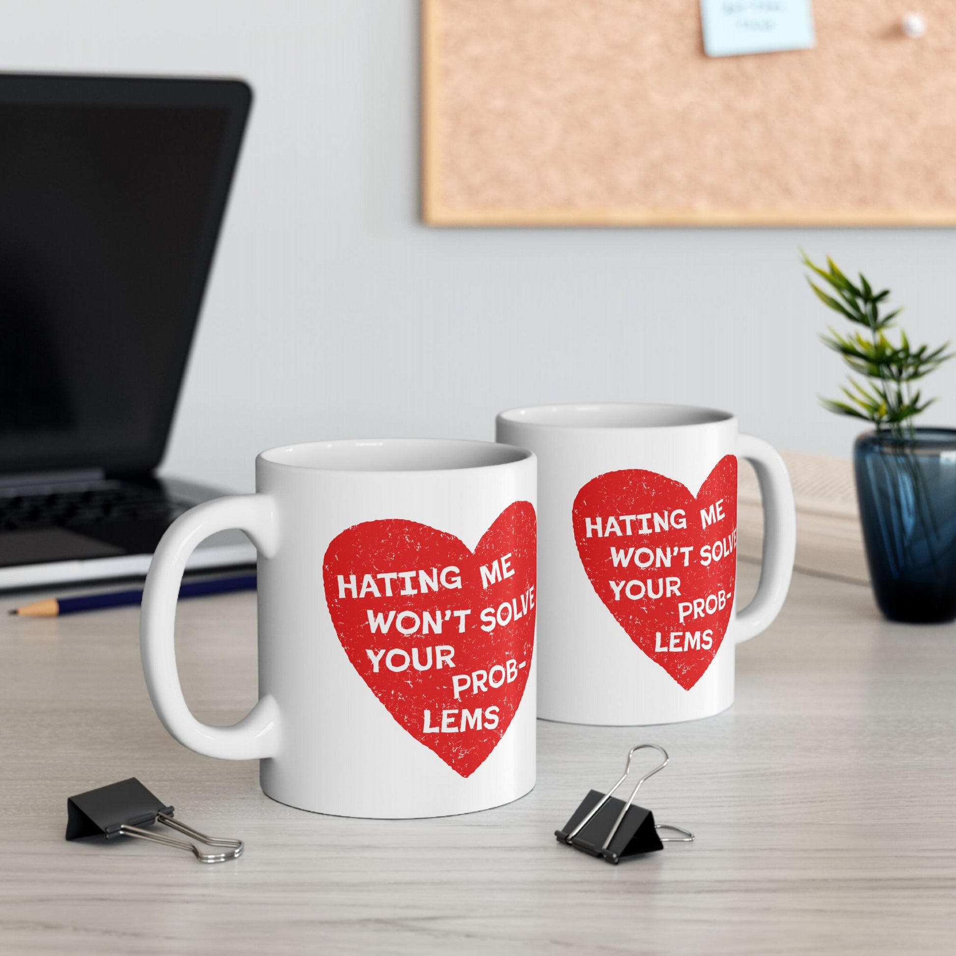 Two white ceramic coffee mugs with a distressed red heart and the slogan “HATING ME WON’T SOLVE YOUR PROBLEMS,” surrounded by stationery, a laptop edge, and a potted plant.