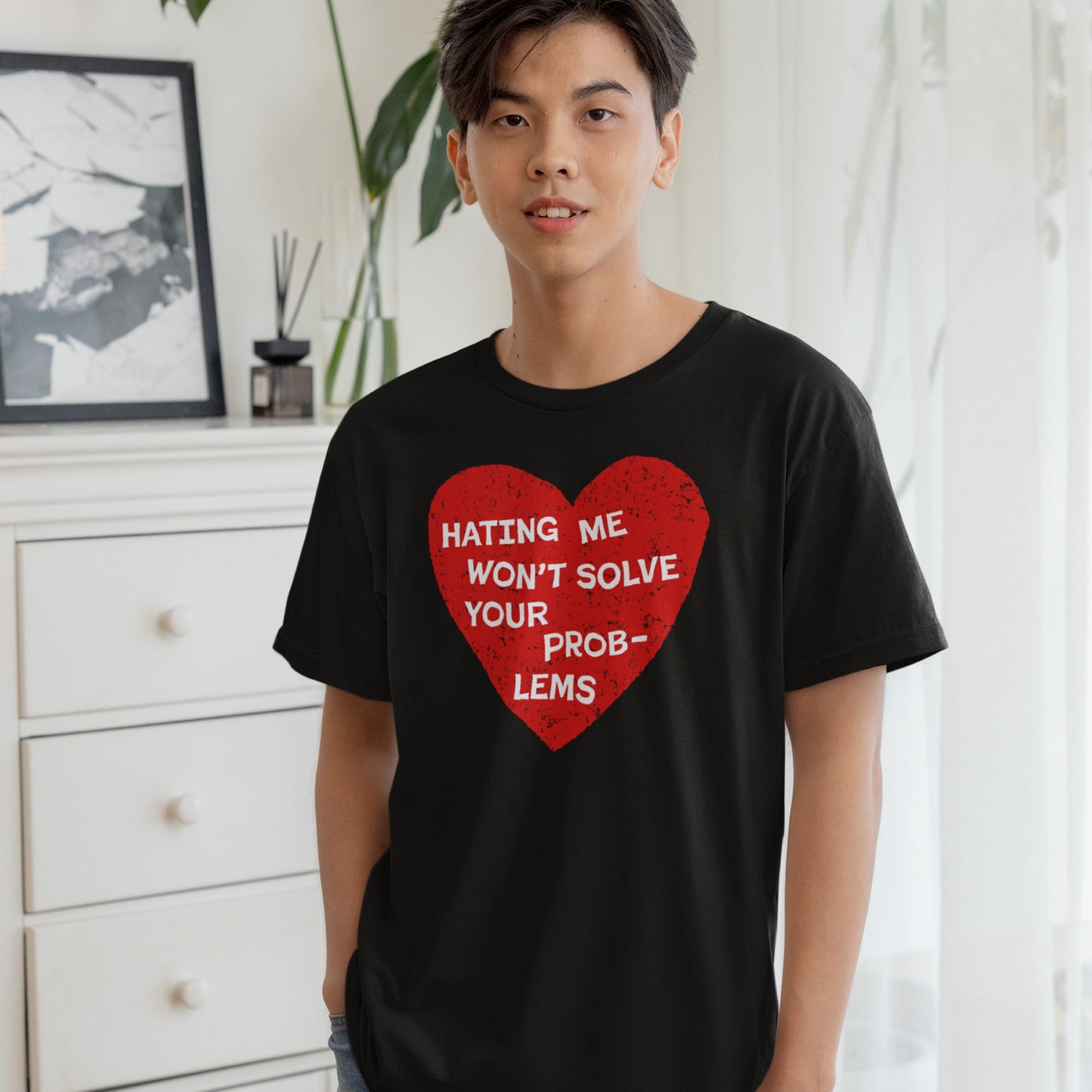 Young man wearing the No Hate T-shirt with a bold red heart graphic and the message “HATING ME WON’T SOLVE YOUR PROBLEMS,” standing indoors near a white dresser and potted plant.