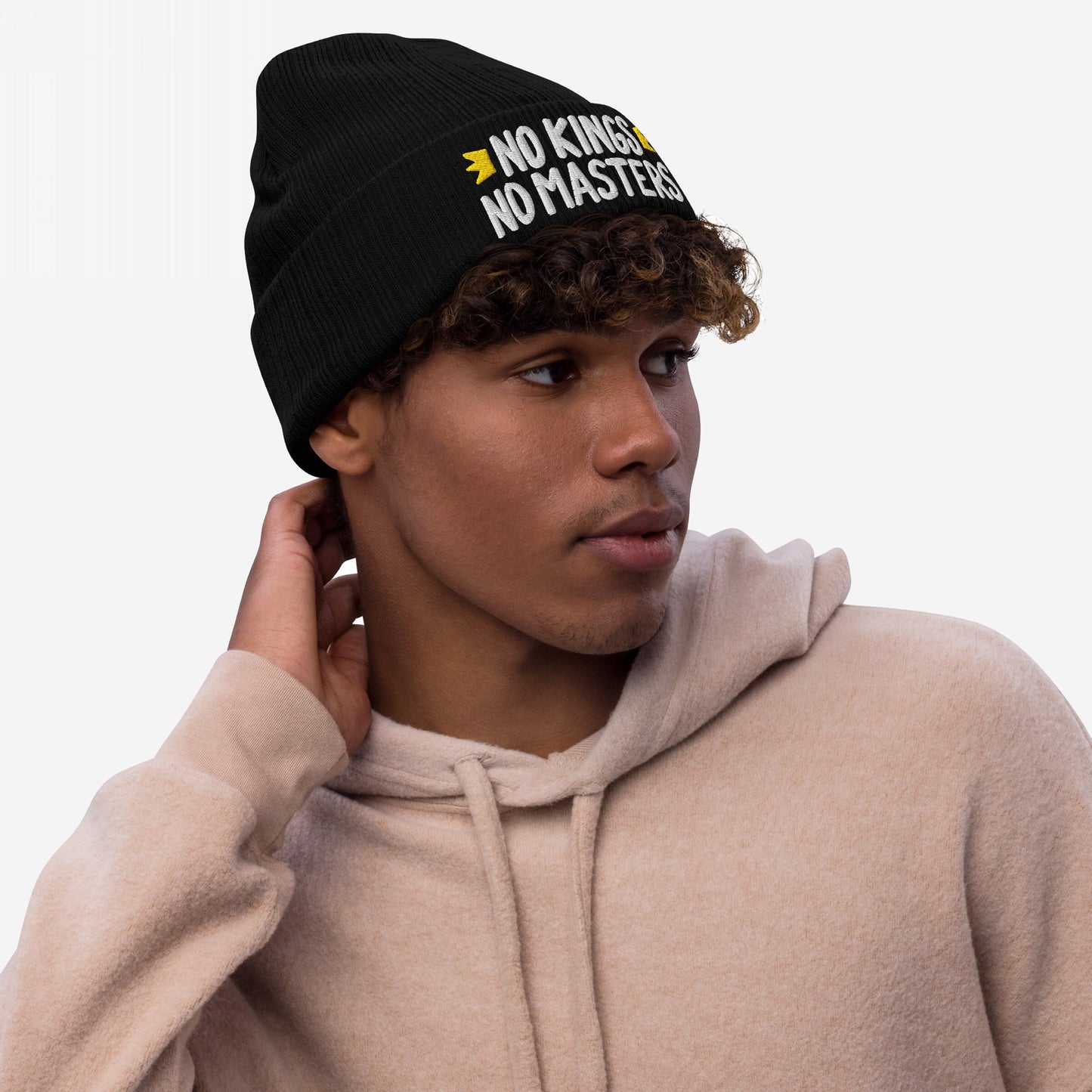 Young man gazing right in a black knit beanie with white NO KINGS NO MASTERS embroidery, featuring yellow arrows, embodying rebellious spirit and activism.