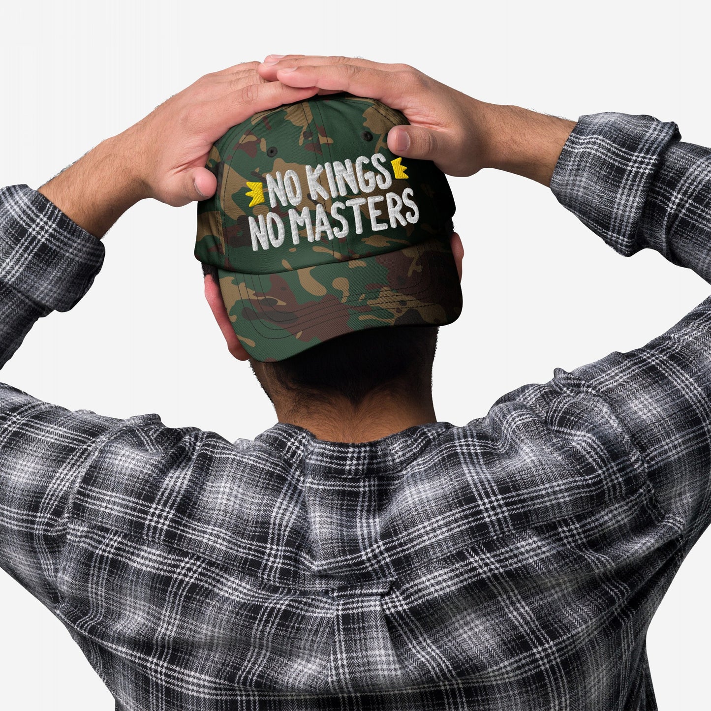 Person wearing the No Kings No Masters Hat, a green camouflage cap with an embroidered slogan, paired with a black-and-white plaid shirt. Hands rest atop the cap, reinforcing a rebellious statement.