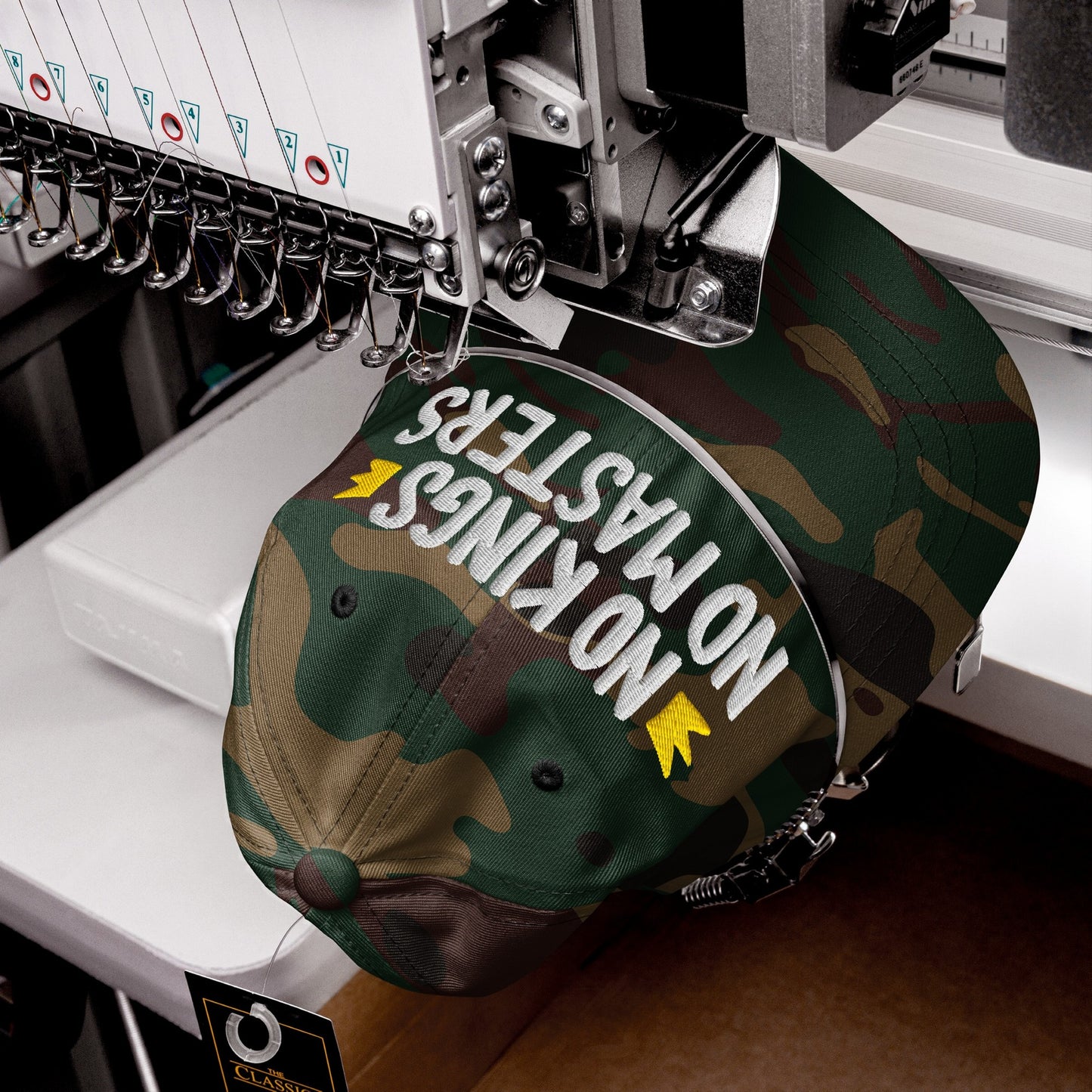 Industrial embroidery machine stitching No Kings No Masters on a camouflage baseball cap. Features raised white thread and yellow accents. CLASSIC tag attached, embodying rebellion and style.