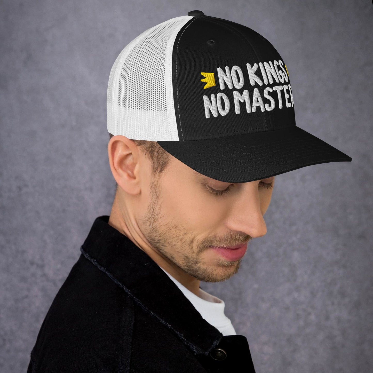 Young man in profile wearing the No Kings No Masters black-and-white mesh trucker cap with bold embroidered message, embodying rebellion and style. Casual attire, gray backdrop highlights the cap's striking design.