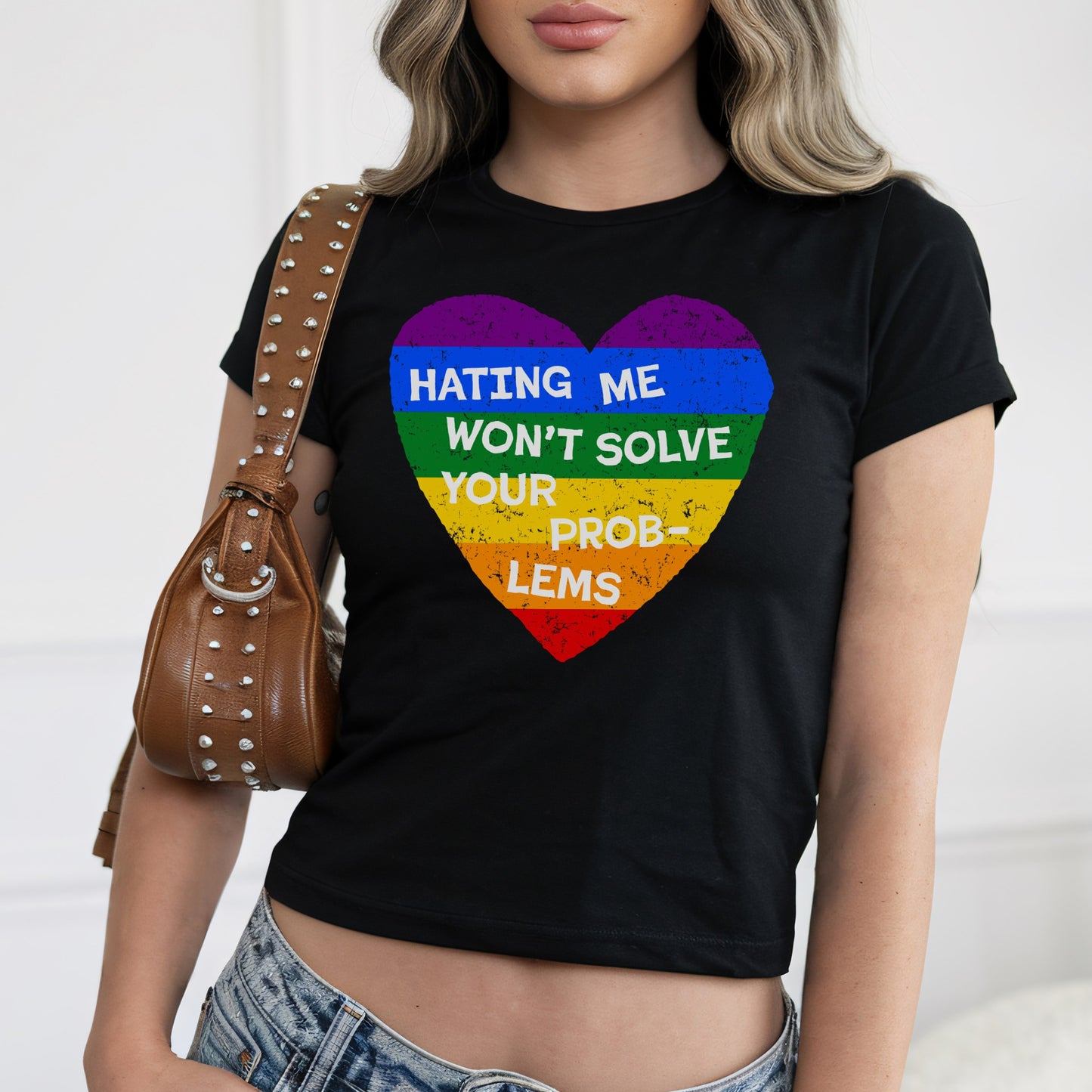 No LGBTQ Hate Women's Baby Tee featuring a rainbow-striped heart with the message “Hating me won’t solve your problems.” Paired with a studded shoulder bag and light-wash jeans.
