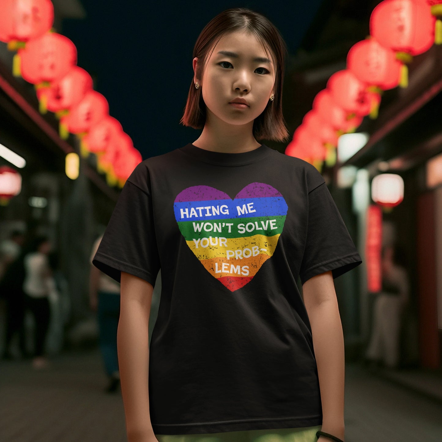 Young person wearing the No LGBTQ Hate T-shirt, featuring a rainbow-striped heart with the slogan “HATING ME WON’T SOLVE YOUR PROBLEMS,” standing on a lantern-lit street at night.