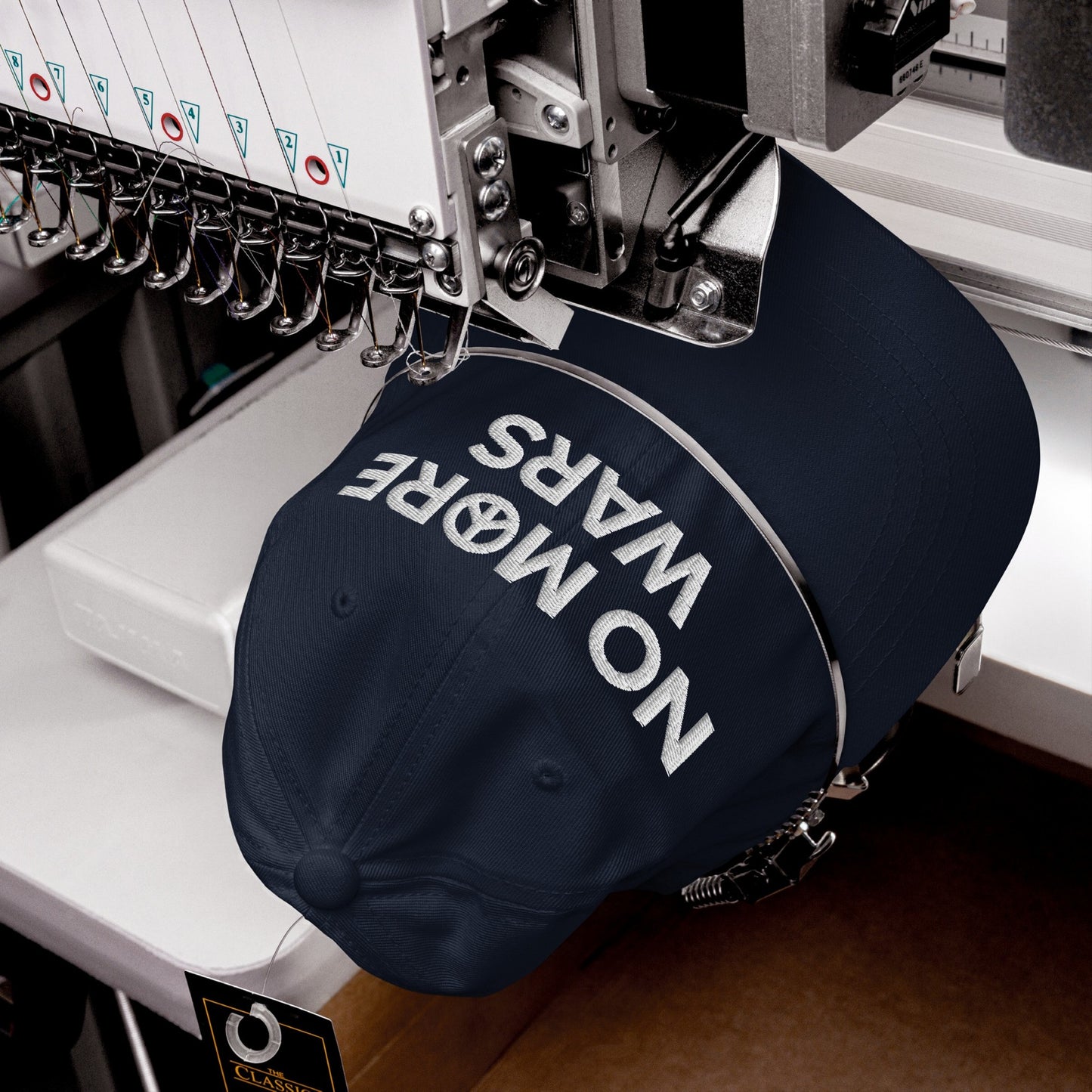 A dark navy No More Wars Hat is secured in an embroidery machine, showcasing its bold NO MORE WARS message with a peace symbol, ready for customization.