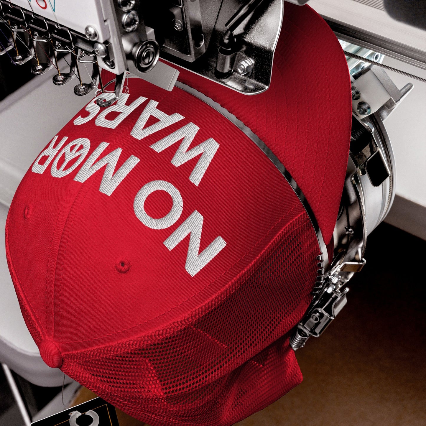 No More Wars Hat being embroidered with the slogan NO MORE WARS on a red trucker cap, secured in an industrial machine, symbolizing a strong anti-war message.