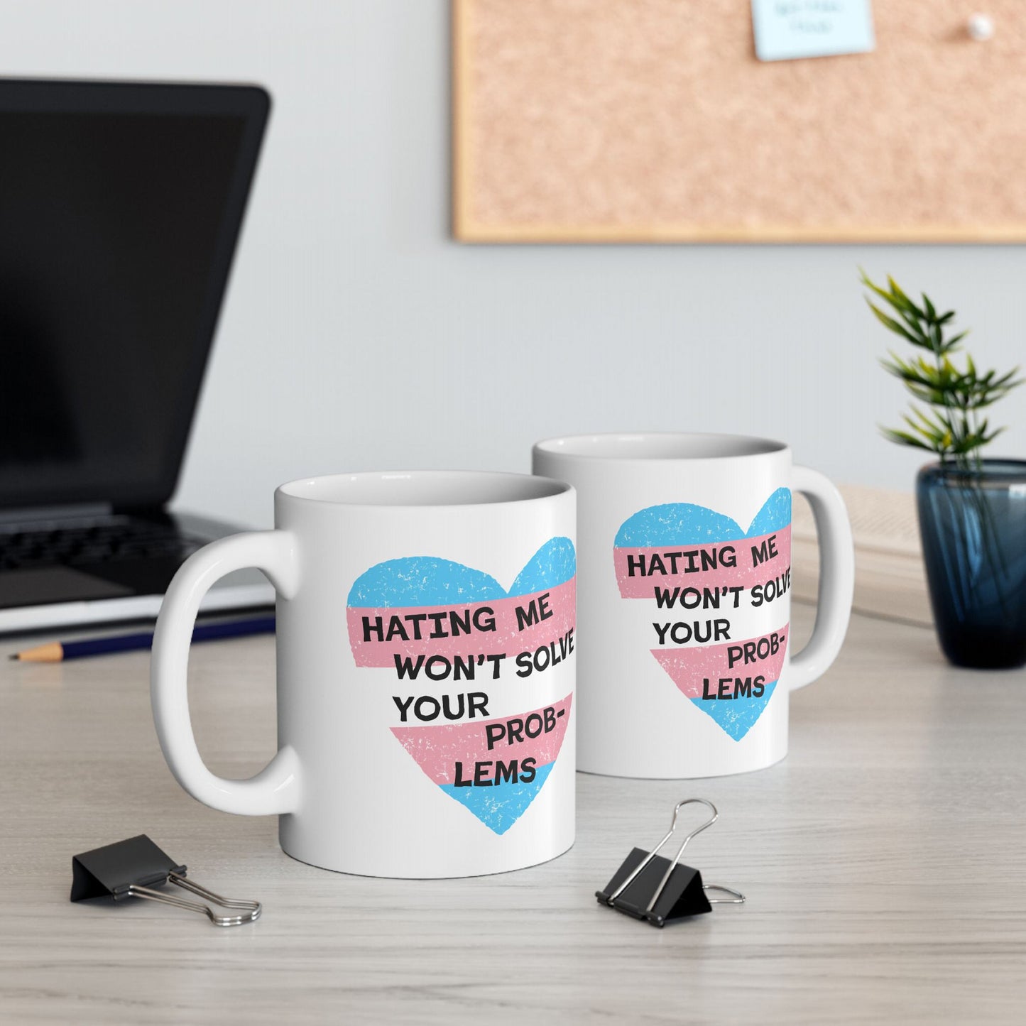 Two white ceramic mugs, each featuring a distressed heart with trans flag colors and bold text: No Trans Hate Mug: Hating Me Won’t Solve Your Problems, on a light wood desk.