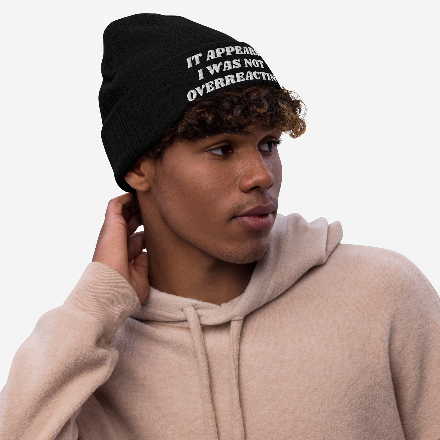 Young man in beige hoodie wearing the Overreacting Hat—a black knit beanie with IT APPEARS I WAS NOT OVERREACTING embroidered in white, embodying activism and resilience.