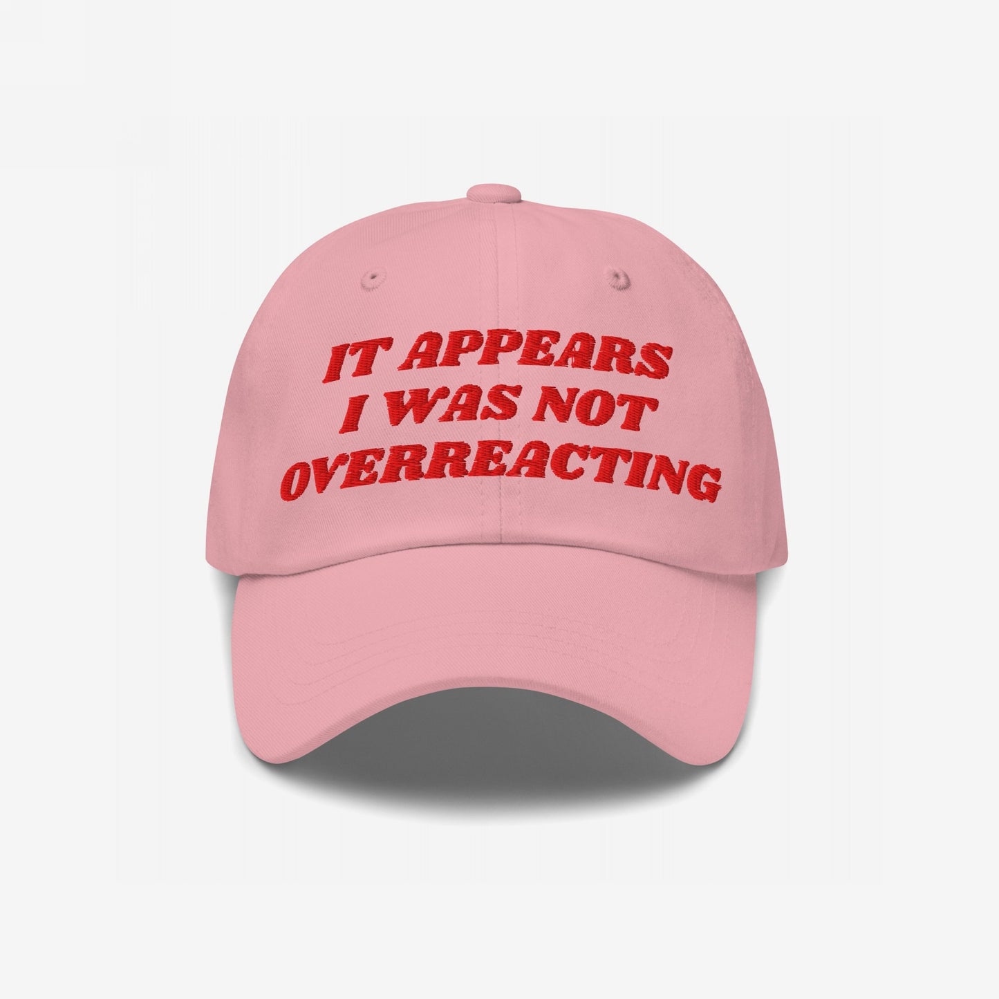 Pink baseball-style cap with red embroidered text, IT APPEARS I WAS NOT OVERREACTING, embodies the Overreacting Hat, designed for vocal progressives opposing political gaslighting. Available in various styles supporting Amnesty International.