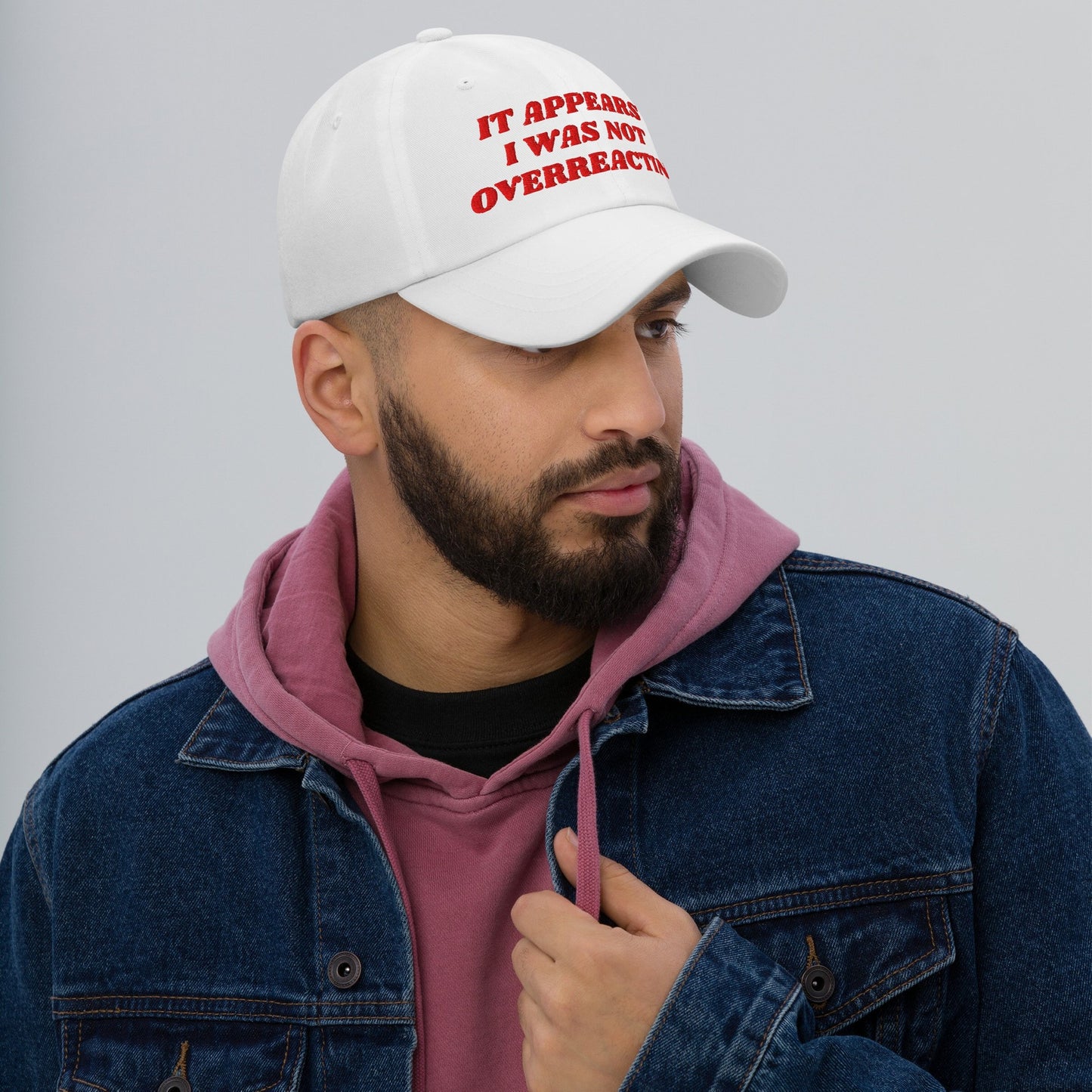 Bearded man models the Overreacting Hat, featuring bold red embroidery on a white cap. Ideal for progressive activists challenging political gaslighting, embodying resilience and advocacy in its design.