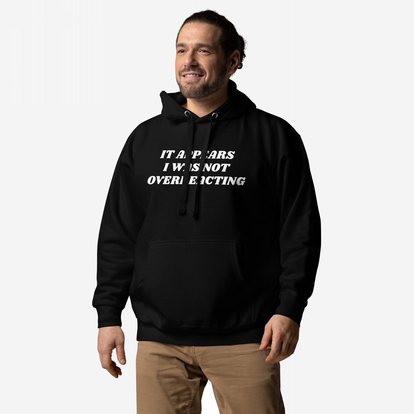 Man in Overreacting Hoodie & Sweatshirt, featuring bold white text critiquing fascism, stands confidently. The black hoodie has a front pocket and drawstrings, paired with khaki pants.
