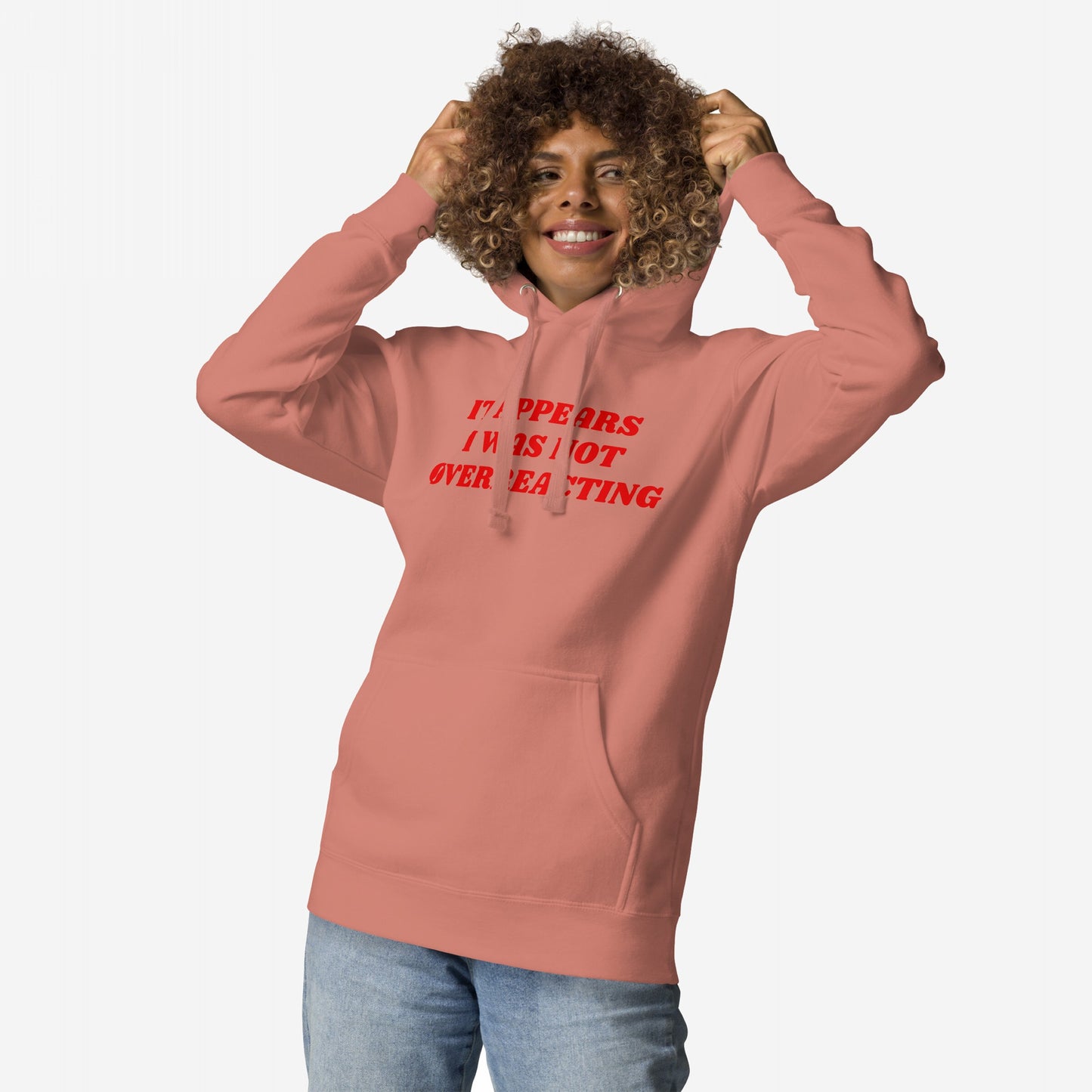 Person wearing the Overreacting Hoodie, pulling its hood up. The hoodie features bold red text, IT APPEARS I WAS NOT OVERREACTING, symbolizing resistance and activism, paired with light-wash jeans.