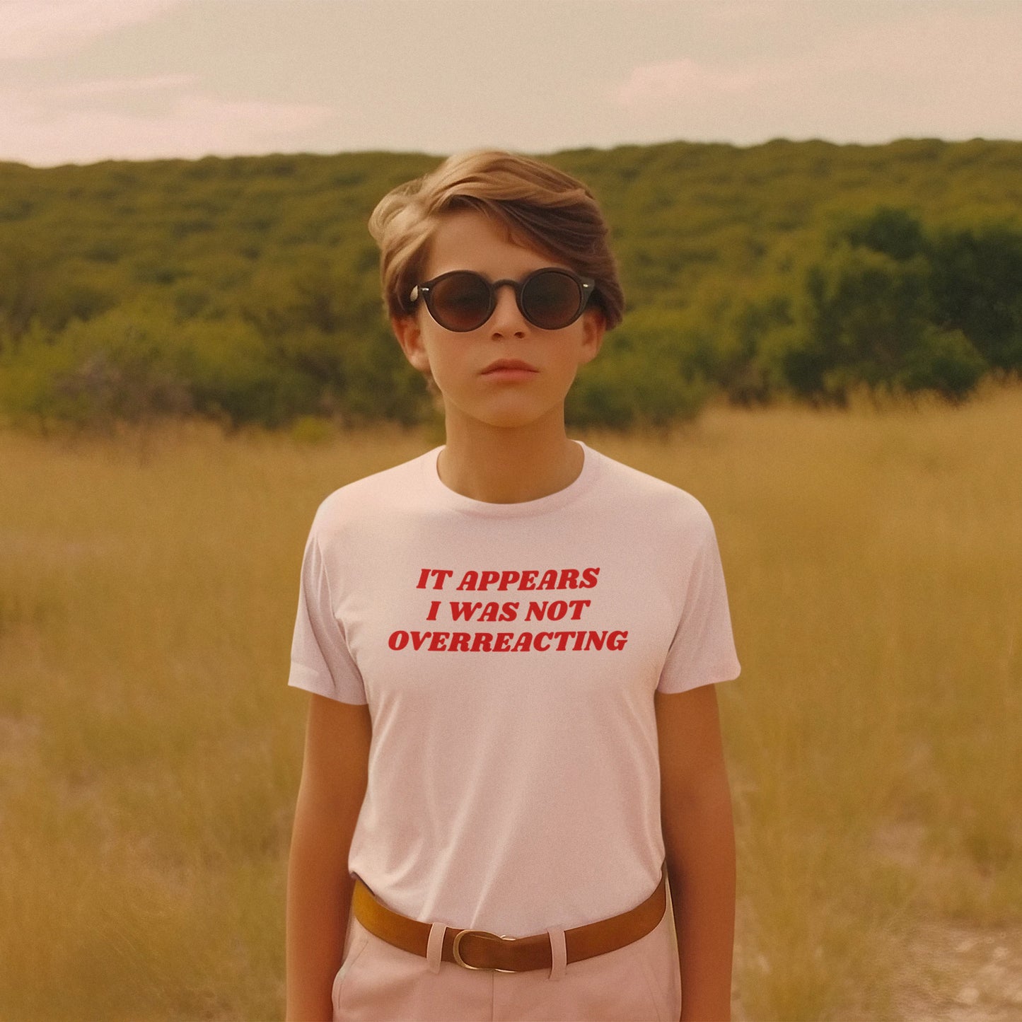 Young person in a grassy landscape wearing the Overreacting Shirt, featuring bold red text: “IT APPEARS I WAS NOT OVERREACTING,” tucked into light trousers, embodying a stance against fascism.