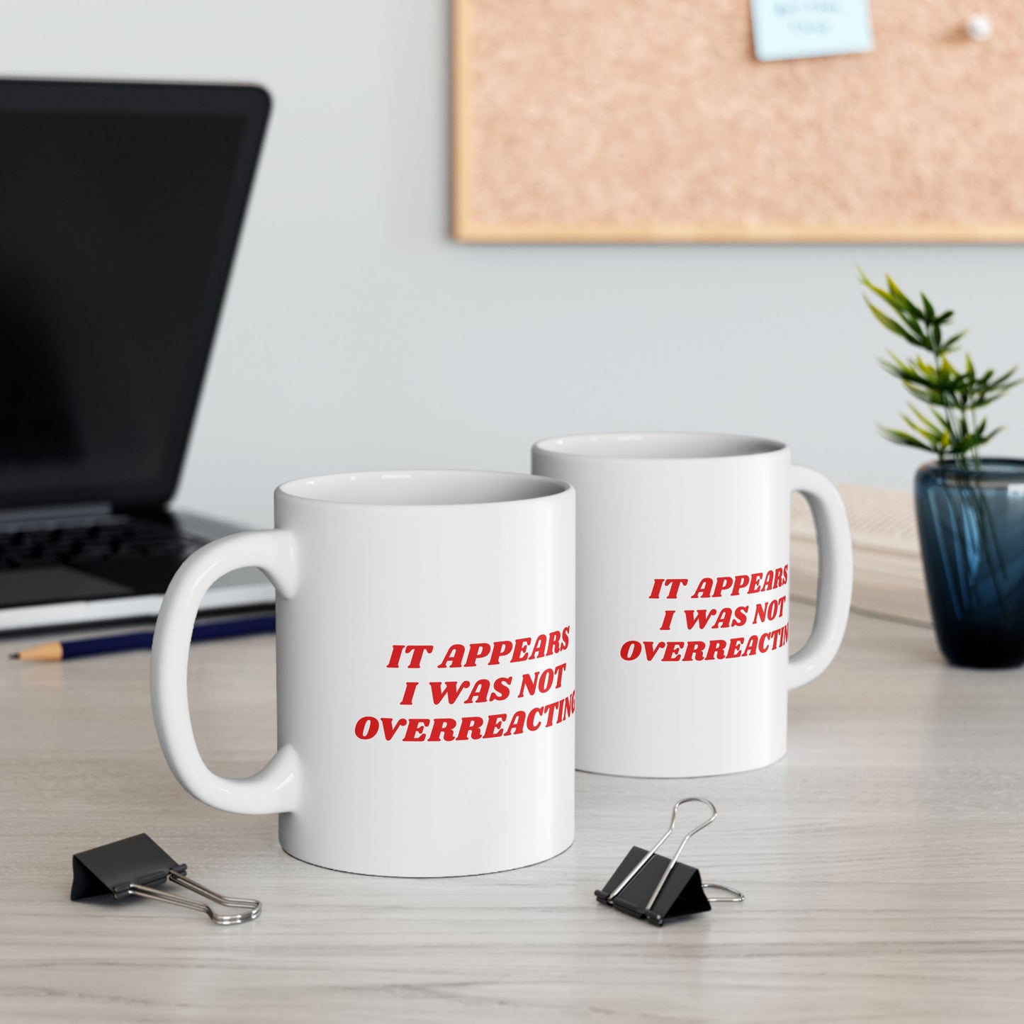 Two white ceramic Overreacting Mugs with red slogan on a desk, surrounded by a laptop, pencil, binder clips, plant, and notebook, embodying bold, progressive statements against fascism.